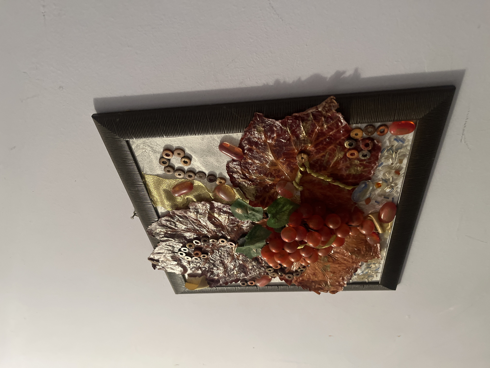 A framed artwork depicting grapes and leaves, possibly a mosaic or mixed-media piece, is mounted on a white ceiling. The frame is dark, possibly black or a dark gray, and appears to have a textured or ribbed edge. The artwork itself features a bunch of red/orange grapes, along with other leaves that are red, green, and textured. Other pieces include brown beads or small objects in patterns, gold fabric and stones. The picture is not in Goce Delchev, Bulgaria.