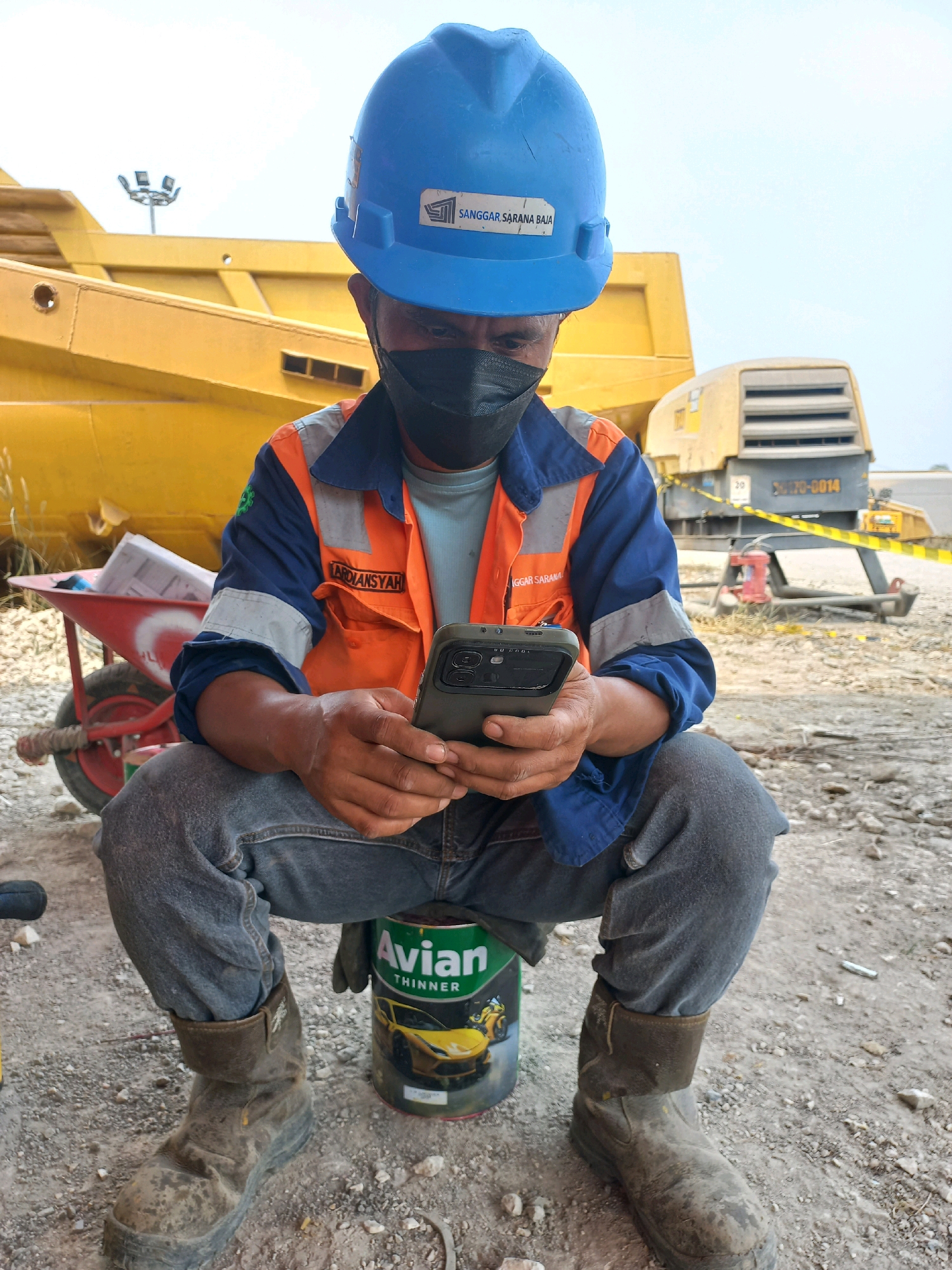 A construction worker in Paringin, Indonesia, wearing a blue hard hat and an orange safety vest over a blue long-sleeved shirt, sits on a can of Avian Thinner. He is looking down at his smartphone, holding it with both hands. The background shows construction equipment, including a large yellow piece of machinery and what appears to be a generator. The ground is dusty and covered with gravel and small rocks. A red wheelbarrow with rolled-up blueprints is positioned to the left of the worker. The worker is also wearing jeans and dirty work boots. The overall scene depicts a break or a moment of rest during a construction project.