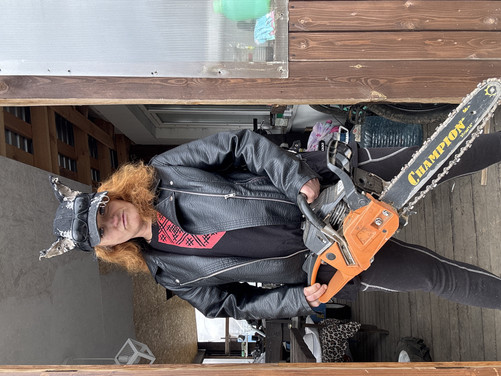 An individual with reddish-blonde, curly hair stands outdoors, holding a chainsaw. They are dressed in a black leather jacket over a black t-shirt featuring a red geometric pattern. A dark cap with cat-like ears and goggles adorns their head, and they wear black leggings with white stitching. The person looks directly at the camera with a neutral expression.
The chainsaw, held by both hands, has an orange engine casing and a black bar. Visible yellow text on the bar reads "CHAMPION" and "35CM/14"". In the background, various objects are discernible, including a large blue plastic bottle with partially visible text "AQUABO," part of a bicycle wheel, and some fabric items, one with a star pattern and another with leopard print. A white metal cage-like object is on the ground to the left.
The setting appears to be a semi-enclosed or open-air structure, possibly a shed or workshop entrance in Chaikovsky, Russia. Overhead, there is corrugated clear plastic sheeting and wooden beams. The ground surface is dark and textured, likely wood or concrete. The lighting is overcast, indicating daytime. The overall scene suggests a practical, outdoor work environment, with the person posing confidently with the heavy-duty tool.