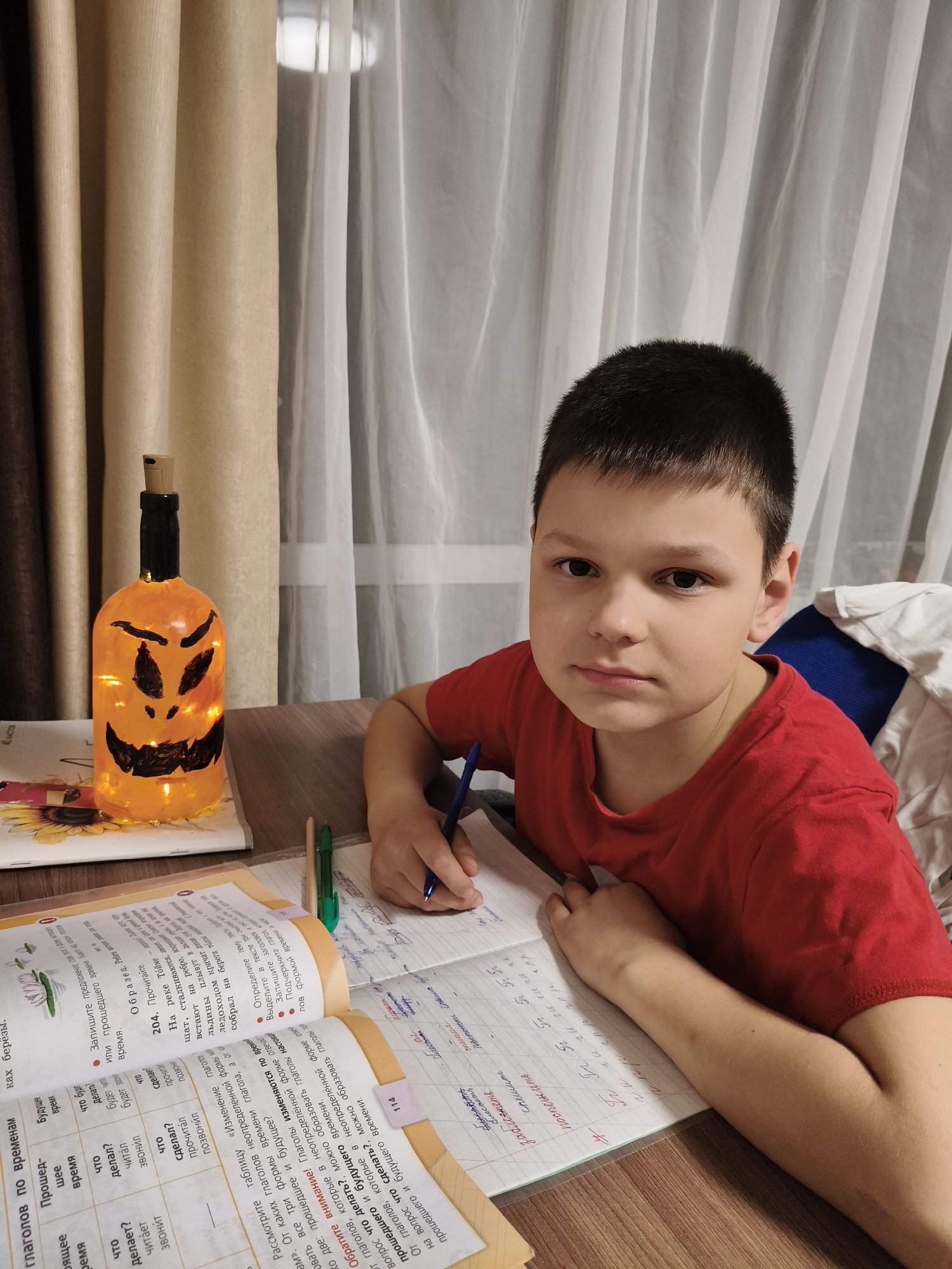 A young boy with short dark hair and wearing a red t-shirt sits at a wooden desk, engrossed in what appears to be homework or studying. He holds a blue pen in his right hand and is writing in an open notebook, while an open textbook lies beside it. The boy looks directly at the camera with a neutral, possibly serious expression.
To his left, a glowing, orange decorative bottle, painted to resemble a jack-o'-lantern with black carved features, sits on the textbook, providing a warm, illuminated ambiance. In the background, there are beige or light brown curtains, partially drawn, with sheer white curtains behind them, revealing a soft light source, possibly a window or another room's light. The setting appears to be indoors, likely a home in the evening, given the artificial light from the bottle and the general lighting of the room.
The textbook is open to pages with Russian text, focusing on grammar. Visible headings include "глаголов по временам" (verbs by tenses), and columns for "Прошедшее время" (Past tense), "Настоящее время" (Present tense), and "Будущее время" (Future tense). Example verbs like "читает" (reads), "звонит" (calls), "делает" (does), "читал" (read - past), and "сделал" (did - perfective past) are present. Instructions like "Рассмотрите таблицу «Изменение глаголов по временам»" (Examine the table "Conjugation of verbs by tenses") and notes such as "Обратите внимание! Глаголы изменяются по временам" (Pay attention! Verbs change by tenses) are clearly legible. The boy's notebook shows handwritten Russian text and numbers, indicating active learning. The scene portrays a focused moment of study in a comfortable, home environment in Chaikovsky, Russia.