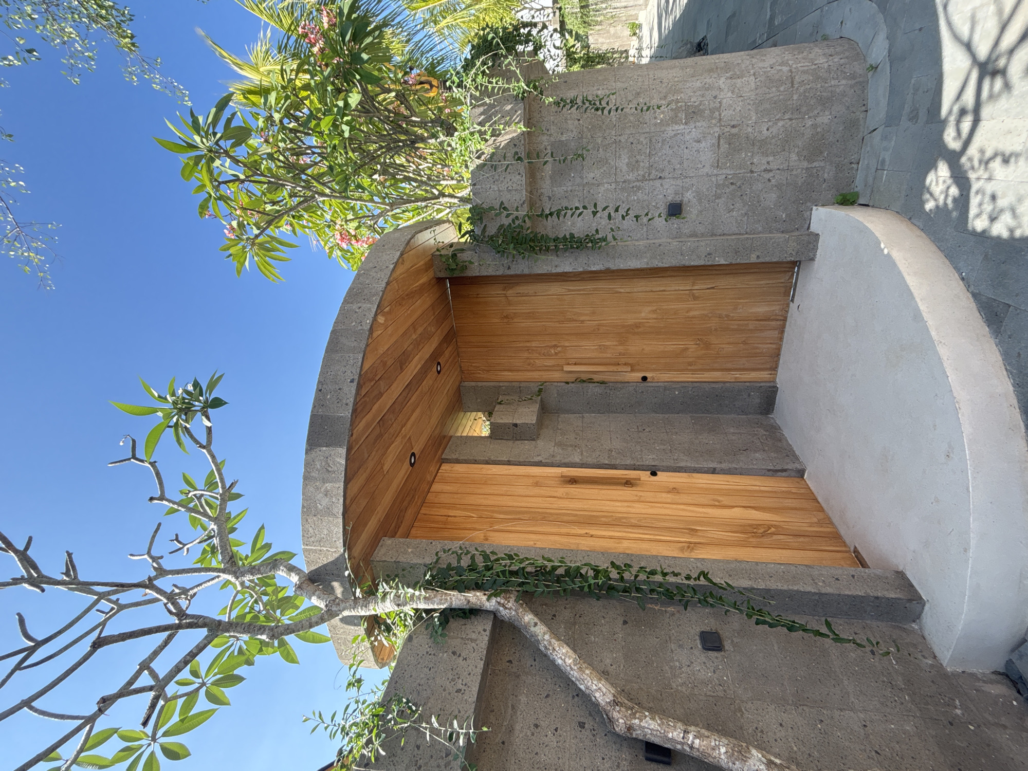 A double wooden door is set into a textured, grey stone wall that curves to the right. The doors are light brown wood with vertical planks and subtle vertical handles. The wall is topped with a curved wooden overhang that mirrors the door's shape. A light-colored, curved concrete step leads up to the doors. Green vines hang from the wall and a tree with bare branches and some green leaves grows from the left side of the wall, its trunk angled across the stonework. Lush green foliage and pink flowers are visible behind the wall on the right. The sky is bright blue. The setting appears to be an entrance to a building or property in a tropical location, likely a resort or private residence in North Kuta, Indonesia, given the vegetation and architectural style.