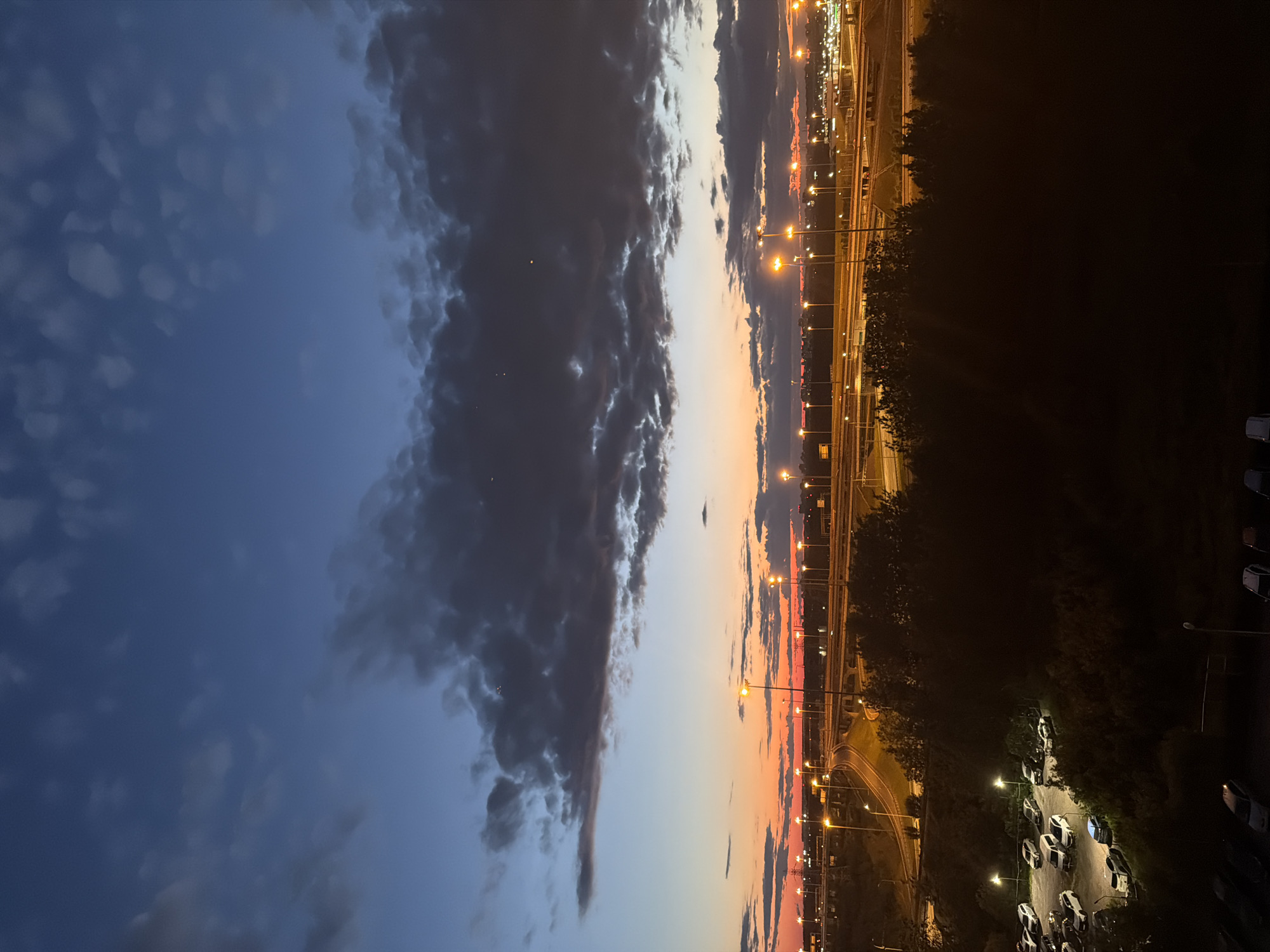 The image depicts a twilight scene overlooking a highway interchange and a parking lot, in an unidentified urban or suburban location. The sky transitions from a deep blue at the top to a vibrant orange and pink gradient near the horizon, indicating either sunrise or sunset. Dark, scattered clouds are present in the upper portion of the sky. Numerous streetlights illuminate the multi-level highway infrastructure, with cars visible on the roadways. In the foreground, a dimly lit parking lot contains several parked cars. A dense line of trees separates the parking lot from the highway. The overall impression is one of a busy transportation hub at the beginning or end of the day.