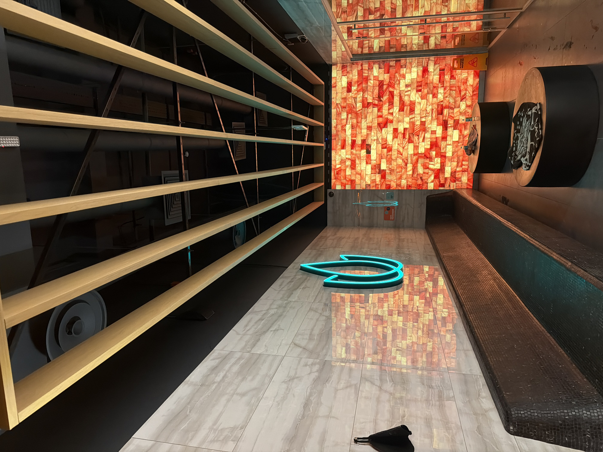 The image shows the interior of a modern spa or relaxation area. The ceiling features a black expanse with a slatted wooden structure running lengthwise, interspersed with exposed pipes and lighting fixtures. The walls are a mix of light-colored, marble-patterned tiles and a striking wall of illuminated Himalayan salt blocks, casting a warm, orange-red glow. A dark, mosaic-tiled bench runs along the left side of the room, with a neon blue flame-shaped sign mounted on the tiled wall above it. In the center of the room, two dark, circular platforms serve as tables, each holding a towel and a bottle of water. To the right, glass doors suggest a separate room or enclosure, also lined with the glowing salt blocks. The floor is a dark, reflective material, possibly polished concrete. The overall ambiance is serene and inviting, with a focus on warm lighting and natural textures.