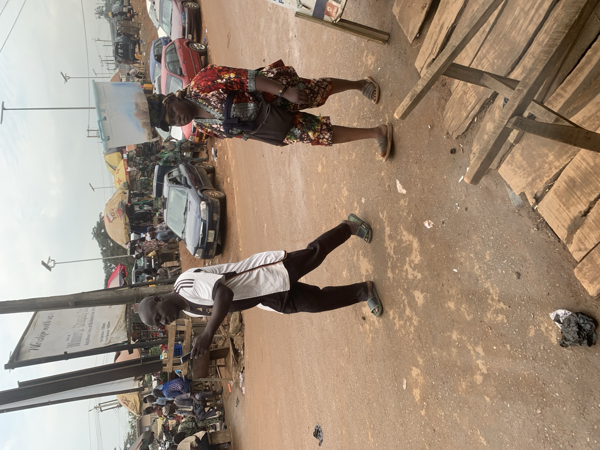 In Shagamu, Nigeria, a man in a white and black shirt and black pants walks while looking at his phone. He wears green sandals. To his right, a woman with a colorful patterned outfit and a black fanny pack walks with a large clear plastic container on her head. She wears brown sandals. The ground is dirt, and there are several cars and people in the background. Umbrellas are set up, suggesting a market or roadside gathering. Wooden planks are visible in the foreground.