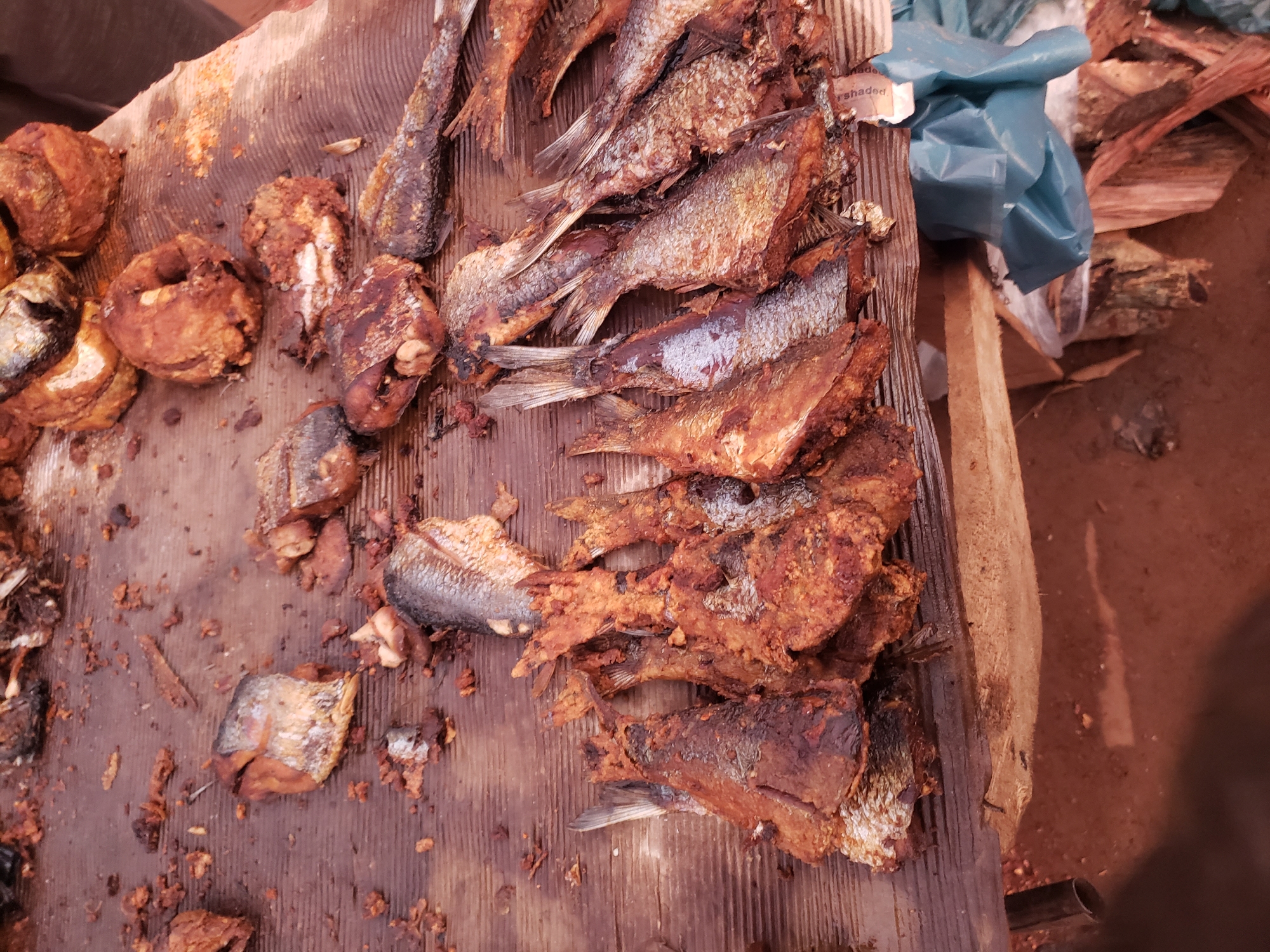 In Kakuri Gwari, Nigeria, a weathered wooden table is laden with an assortment of fried fish. The fish, varying in size and preparation, display a deep golden-brown hue, indicating they are thoroughly cooked. Some are whole, while others are cut into pieces, their scales still visible on some of the larger specimens. Crumbs and small fragments of fried fish are scattered across the table's surface, suggesting a bustling market environment. In the background, portions of what appear to be more fried fish or food items are visible. The table is situated outdoors, with a reddish-brown dirt floor visible beneath it. To the right of the table, a blue plastic bag and some stacked pieces of firewood are present, adding to the rustic setting.