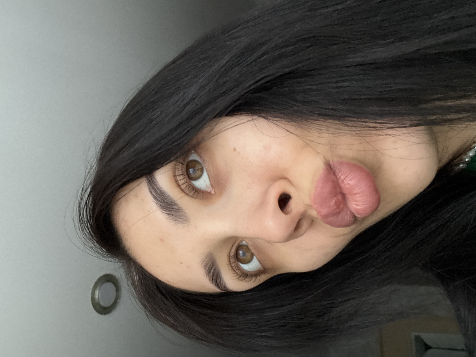 A close-up, rotated image shows a woman with dark hair, brown eyes, and fair skin. She is making a duck-face expression. A light grey wall with a round fixture is visible in the background. No information suggests the location is in Votkinsk, Russia.