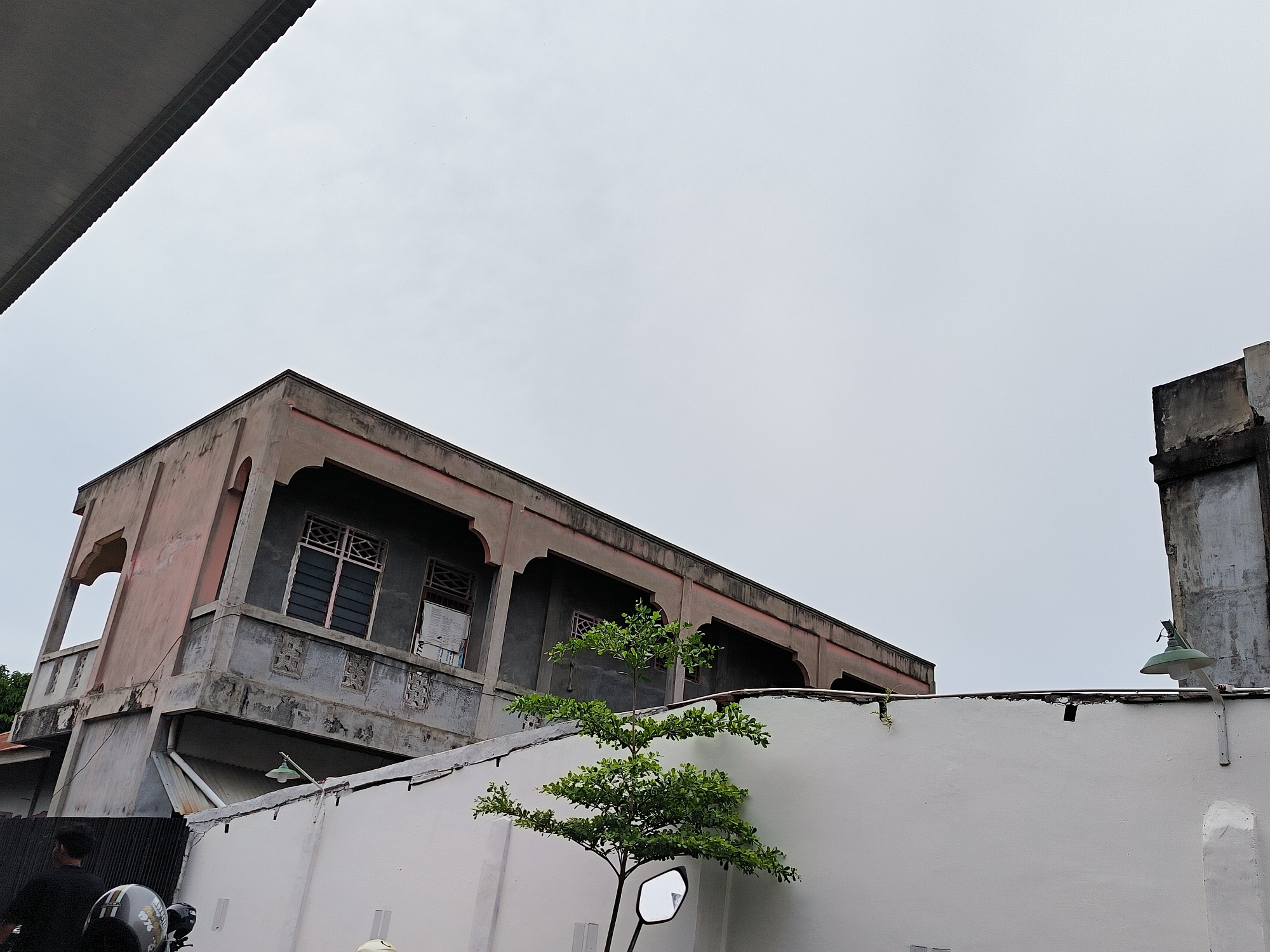 A low-angle shot reveals a two-story building with a weathered pink and grey facade in Lhokseumawe, Indonesia. The building's architecture features arched openings and decorative elements beneath the windows. Adjacent to the building is a white wall with a small green tree in front. A street light is mounted on the wall. The sky is overcast. In the foreground, a motorcycle with a helmeted rider is partially visible.