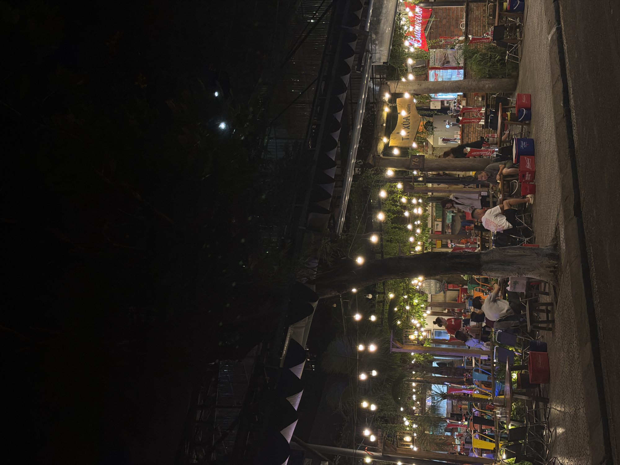 The scene depicts a lively street-side establishment in Đà Nẵng, Vietnam at night. String lights illuminate a narrow street filled with small tables and chairs occupied by people. Some individuals sit and relax, while others appear to be engaged in conversation or enjoying food and drinks. Beer logos like "Budweiser" are visible, suggesting the venue serves alcoholic beverages. Some customers sit on red beer crates. The surrounding buildings are partially visible, and trees or foliage are incorporated into the decor, giving the place a slightly organic and casual feel.