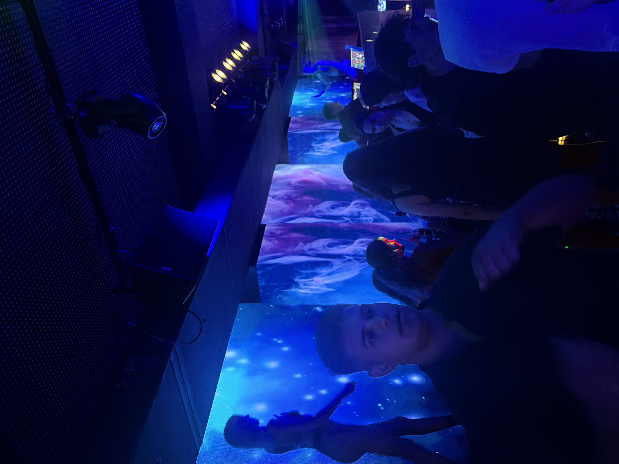 In a nightclub in Sozopol, Bulgaria, patrons are illuminated by blue and purple lights. A man in the foreground, wearing a dark shirt, is looking forward with his hands in motion, suggesting dancing. Behind him, a silhouette of a woman in a bikini poses against a backdrop of animated blue lights with white specks. To the right, another backdrop displays swirling purple and white patterns. In the distance, a DJ booth is visible, with a figure standing behind it. Several other individuals are scattered throughout the dimly lit space, their faces obscured by shadows and the vibrant lighting. The ceiling is adorned with a textured acoustic panel, from which spotlights and audio equipment are suspended.
