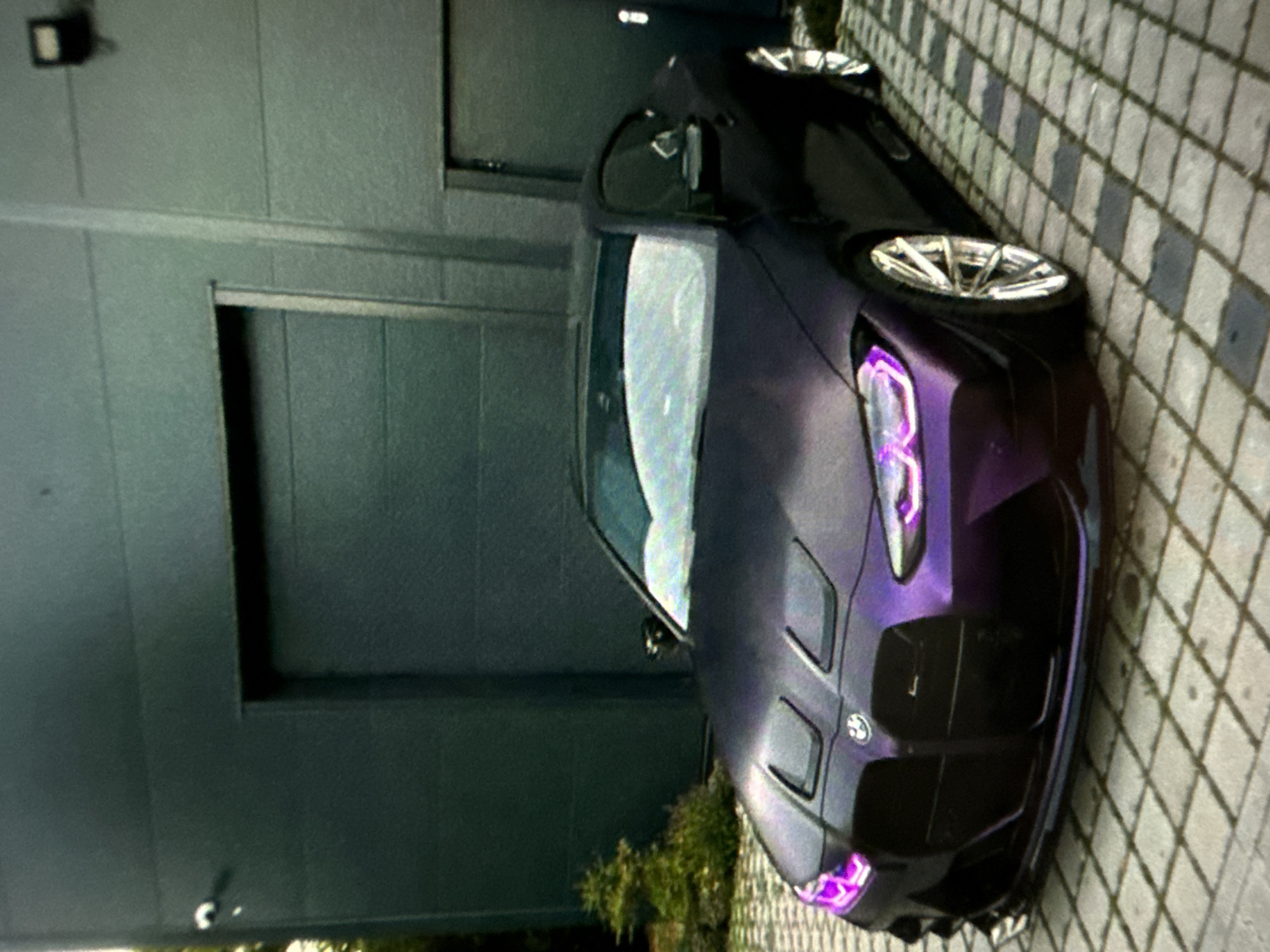 A purple BMW car is parked on a patterned brick surface next to a gray building with a closed garage door. The location appears to be in a city or urban area of Unknown. The car has custom headlights that are illuminated with a purple hue. The scene is static.