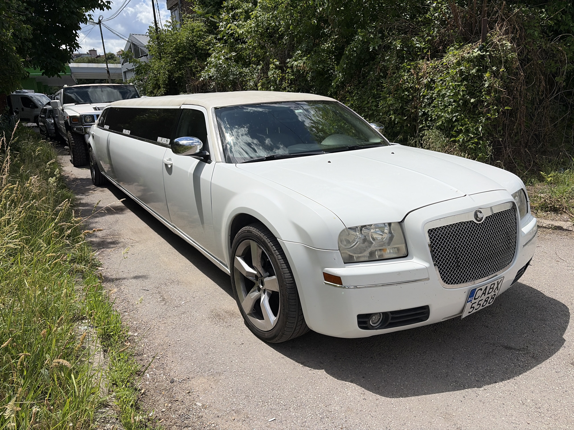 A white Chrysler 300 stretched limousine is parked on an unpaved road in a semi-urban area, likely in Sofia, Bulgaria, given the license plate BG. The limousine is the primary subject, positioned at an angle facing the viewer's right, with its front slightly closer. Its roof is a contrasting light beige or white vinyl. The vehicle's headlights, chrome grille, and front bumper are clearly visible. A Bulgarian license plate with the characters "CABX 5588" is affixed to the front.
Behind the limousine, other vehicles are lined up, including a white Hummer H2 SUV and a dark-colored van. The vehicles are parked along the side of the road, which is bordered by dense, green vegetation and trees. The ground is a mix of dirt and gravel. In the background, glimpses of buildings and utility poles suggest a developed area, possibly a residential or industrial zone on the outskirts of the city. The sky is partly cloudy. The lighting suggests it is daytime.