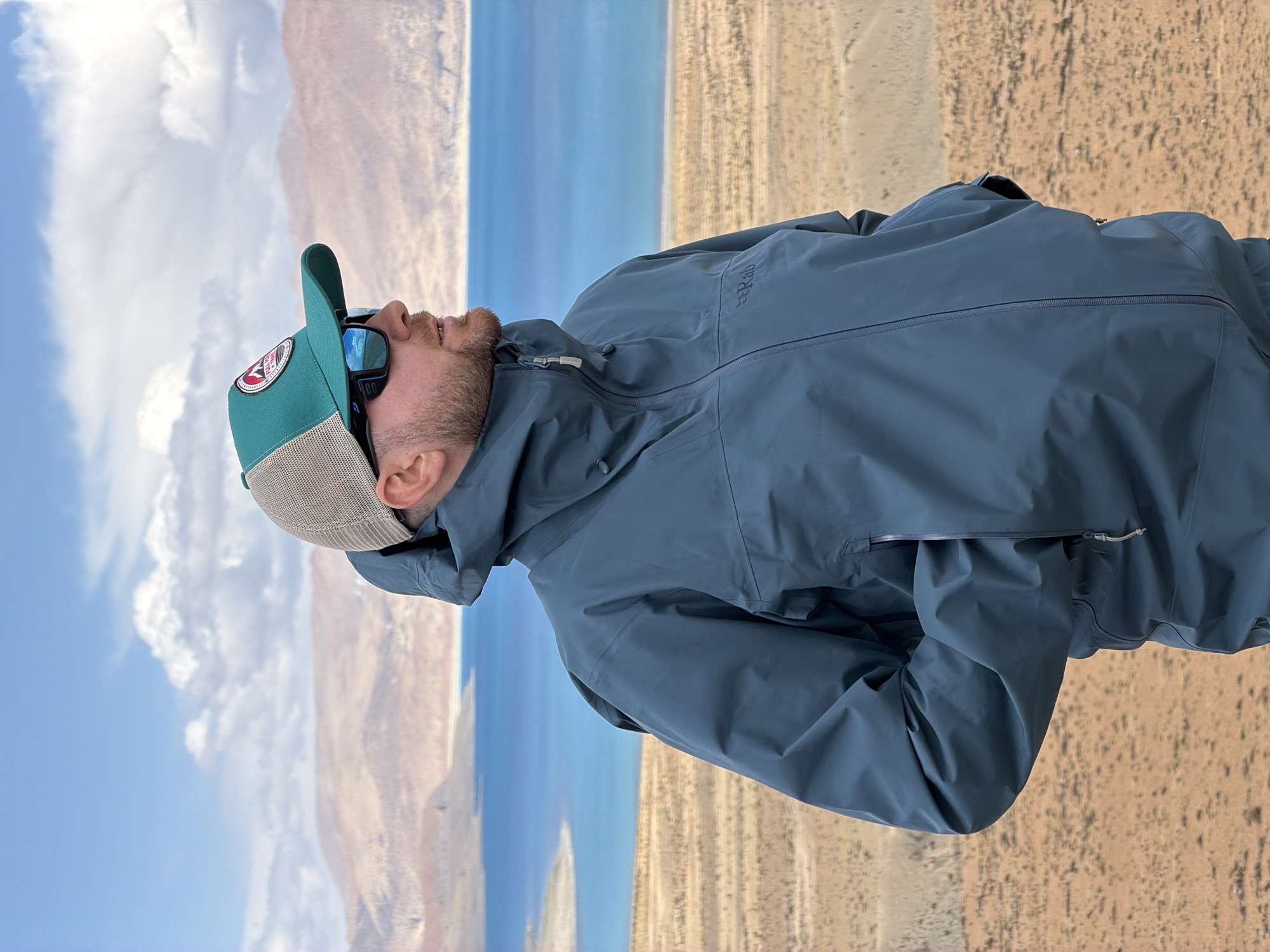 A man stands in an arid landscape near the city of Gyirong County, China. He is wearing a blue-gray waterproof jacket, a green baseball cap with a mesh back, and sunglasses, looking up towards the sky. The landscape includes a blue body of water, likely a lake or river, with sandy-colored terrain on either side. The sky is a mix of blue with white clouds. The man's pose suggests he is observing the sky or the scenery.