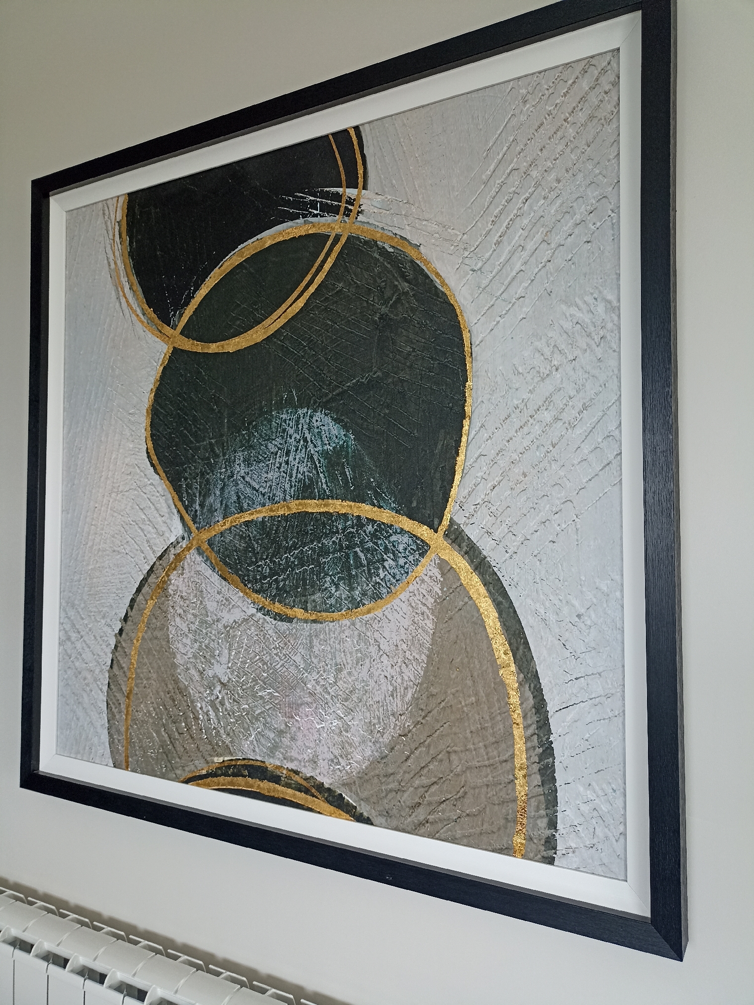 The image shows a framed abstract artwork hanging on a white wall. The artwork features large, overlapping circles in dark green and beige tones, with gold-colored abstract lines overlaying them. The background of the artwork has a textured, rough finish with imprinted lines. The frame is black with a white matting. Below the artwork and slightly to the left, a white radiator is visible. The scene is static and shows no people or actions. The location is specified as Doirí Beaga, Ireland, but there are no visual cues in the image to confirm this location.