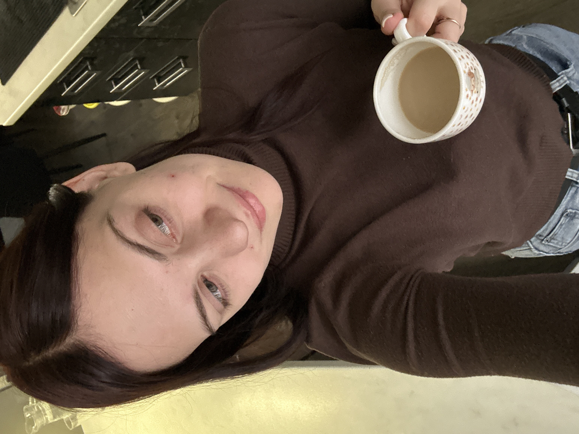 A person is holding a mug in an indoor setting. The person, wearing a brown turtleneck sweater and blue jeans, is holding a white mug containing a light brown liquid. The background includes a white countertop and a section of kitchen appliances. The exact location in Chaikovsky, Russia, cannot be determined from the image.