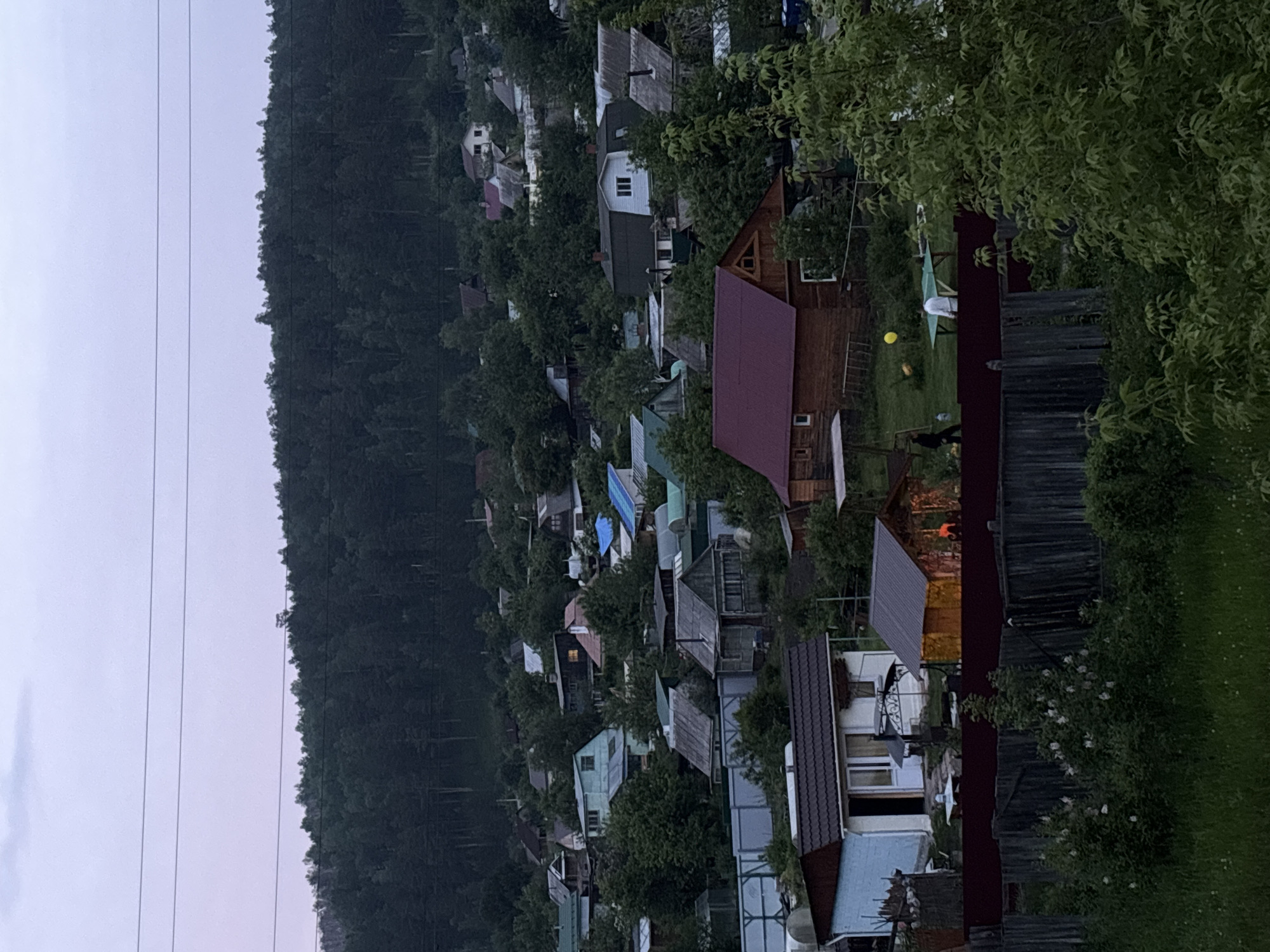 The image shows a view of a dacha settlement (summer cottage community) located on the slopes of a hill covered with a dense forest. The sky is overcast with a pale purple hue. Several houses with different roof colors are visible, surrounded by trees. Some houses have small gardens. In the foreground, a wooden fence is prominent. A ping pong table with two people, one in white and the other in black, are visible in the middle ground. A yellow balloon is also visible in the area. The image captures a moment of leisure activity in a rural setting, likely during summer or early autumn. The settlement appears to be part of the suburban area of Zheleznogorsk, Russia, given the characteristic architecture and landscape.