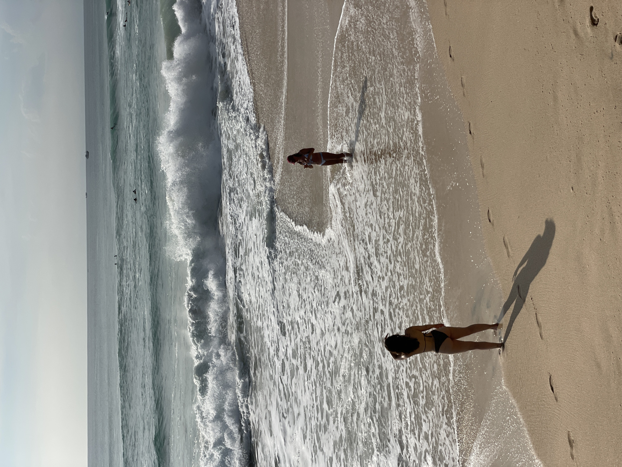 This photograph shows a beach scene in Kuta Selatan, Indonesia. The image is oriented vertically. In the foreground, a woman stands at the edge of the water. Her shadow stretches onto the sand. Further away, another woman is visible, standing in the shallows. The ocean is characterized by crashing waves. The sand is light tan, and the water is a greenish-blue hue. The sky is partially visible at the top left, appearing overcast. There are a few specks in the distance that could potentially be birds flying over the water. Footprints dot the sand near the water's edge.
