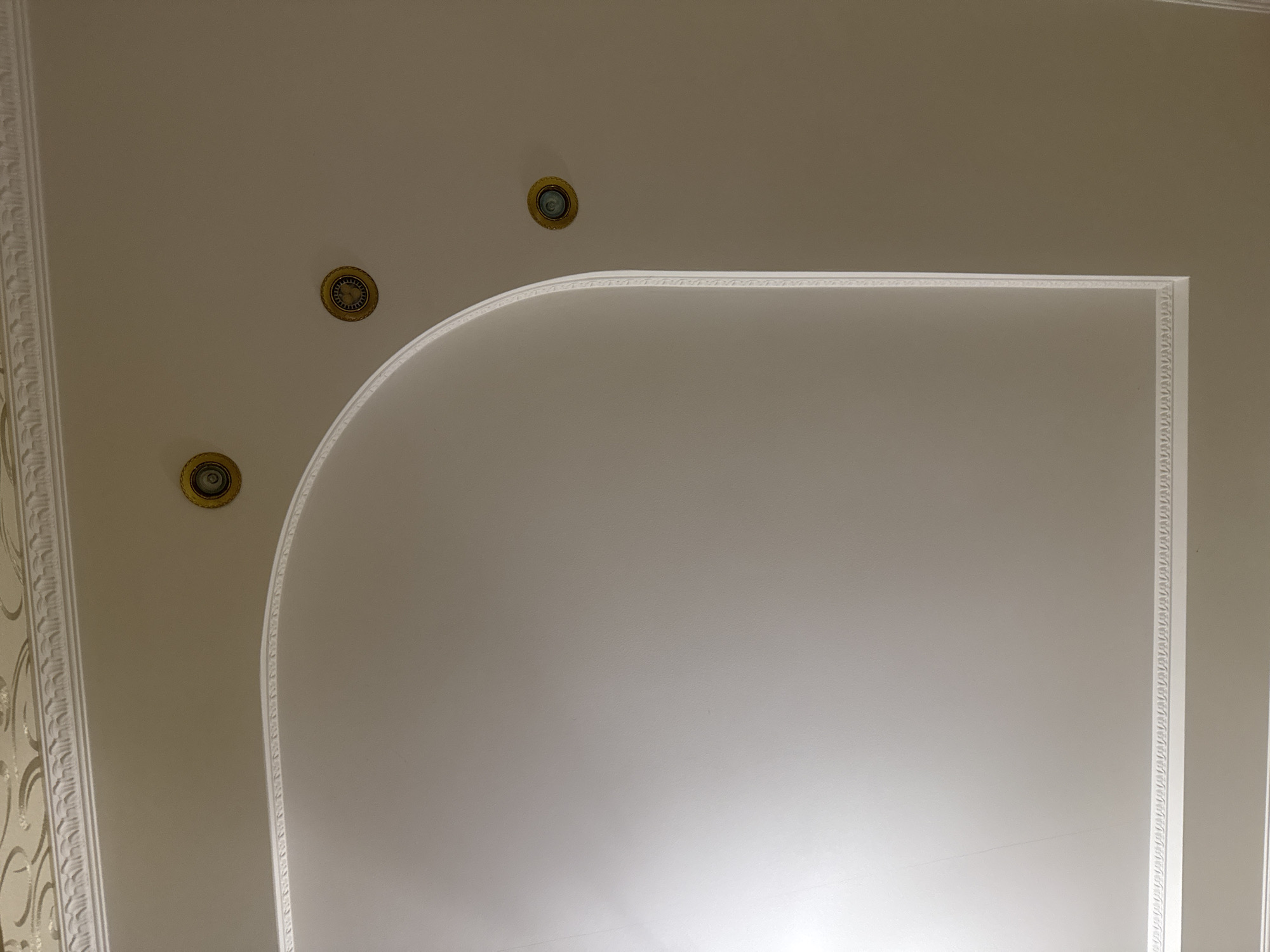 The image shows a section of an interior ceiling with decorative elements. The ceiling is painted a light beige color and features a recessed area with curved edges. This recessed area is outlined with white decorative molding that has a repeating pattern. There are three circular recessed light fixtures, each with a gold-colored trim, embedded in the main ceiling area. Above the recessed section, a decorative frieze runs along the top edge of the image, also in white. The lighting in the room appears to be coming from the recessed lights and possibly from an unseen source, casting soft shadows and highlighting the textures of the ceiling and molding. The overall impression is one of a well-decorated interior space, possibly a living room or dining area. The image does not contain any people or signs of human activity beyond the installed fixtures and decorative features. The location in Гожули, Ukraine, is not discernible from the visual elements of the image itself.
