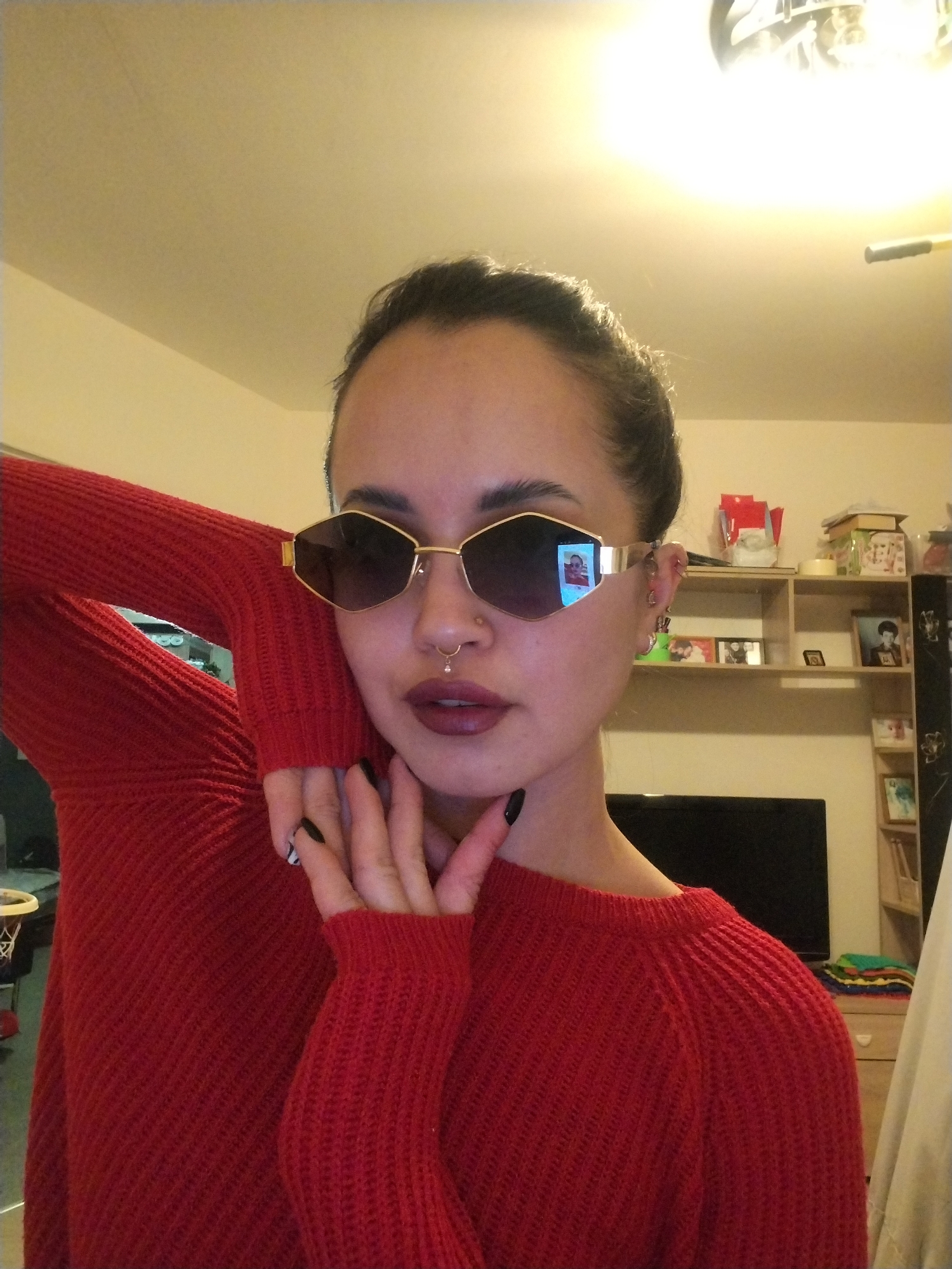 A woman wearing a red knitted sweater and gold-rimmed sunglasses with dark lenses poses indoors. She has her hair pulled back and is wearing dark lipstick and a nose ring. Her left hand is near her face, with fingernails painted black and a design on one nail. Behind her is a shelving unit with various objects, including photographs and decorative items. A television is visible to the right of the shelving unit. The location is inside a home in an unknown city.