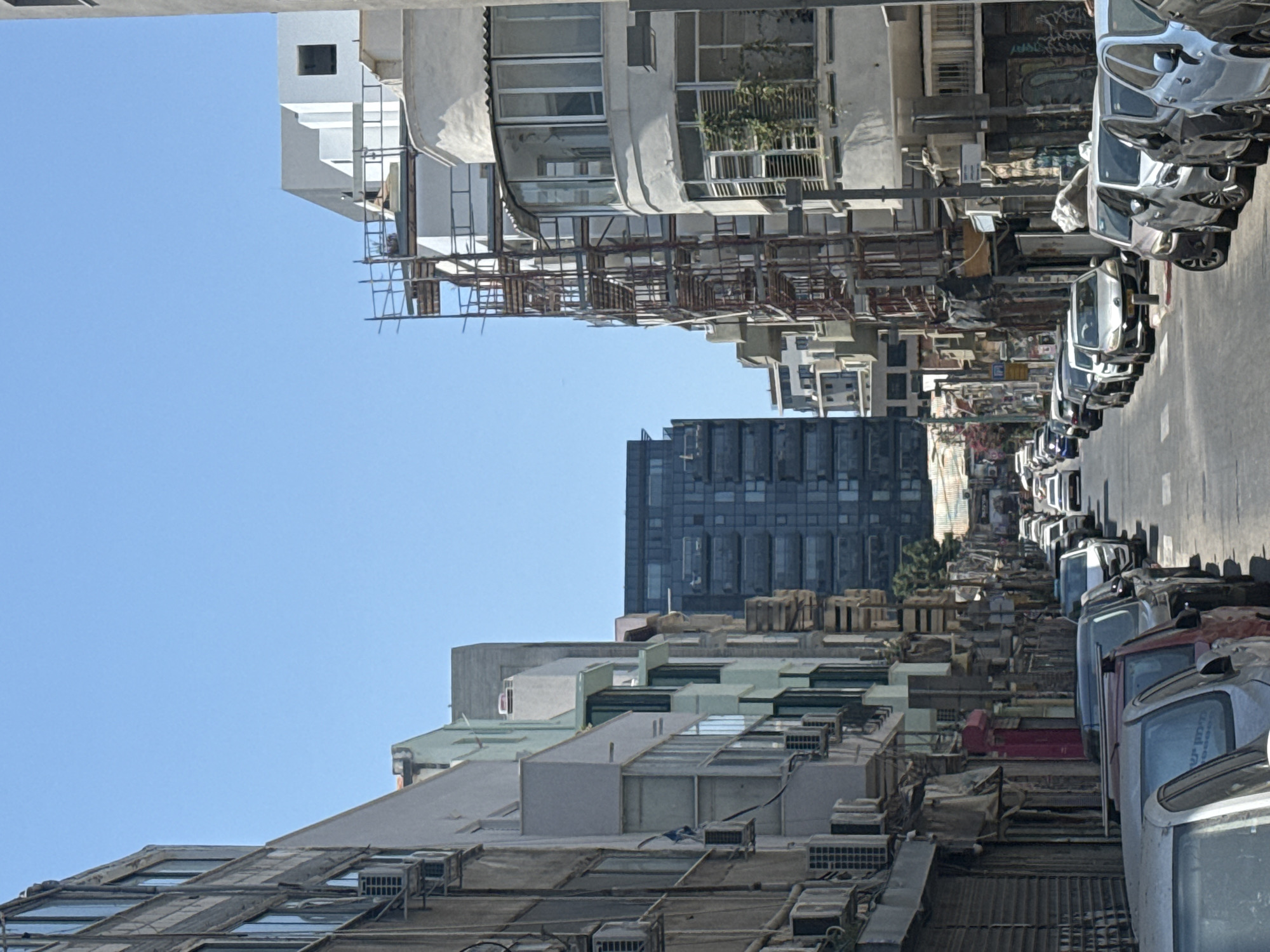 This is a photograph of a street in Tel Aviv, Israel, under a clear blue sky. The perspective is looking down a narrow street lined with multi-story buildings on both sides. The buildings vary in architectural style, with some appearing older and others under construction or renovated, evidenced by scaffolding on the right side. The street is filled with parked cars along both curbs, and a line of cars is visible in the distance, suggesting traffic. The buildings feature numerous windows and air conditioning units. A taller, dark glass building is visible in the background, adding a modern contrast to the surrounding structures. The scene depicts a typical urban environment in Tel Aviv, characterized by its dense architecture and active street life.