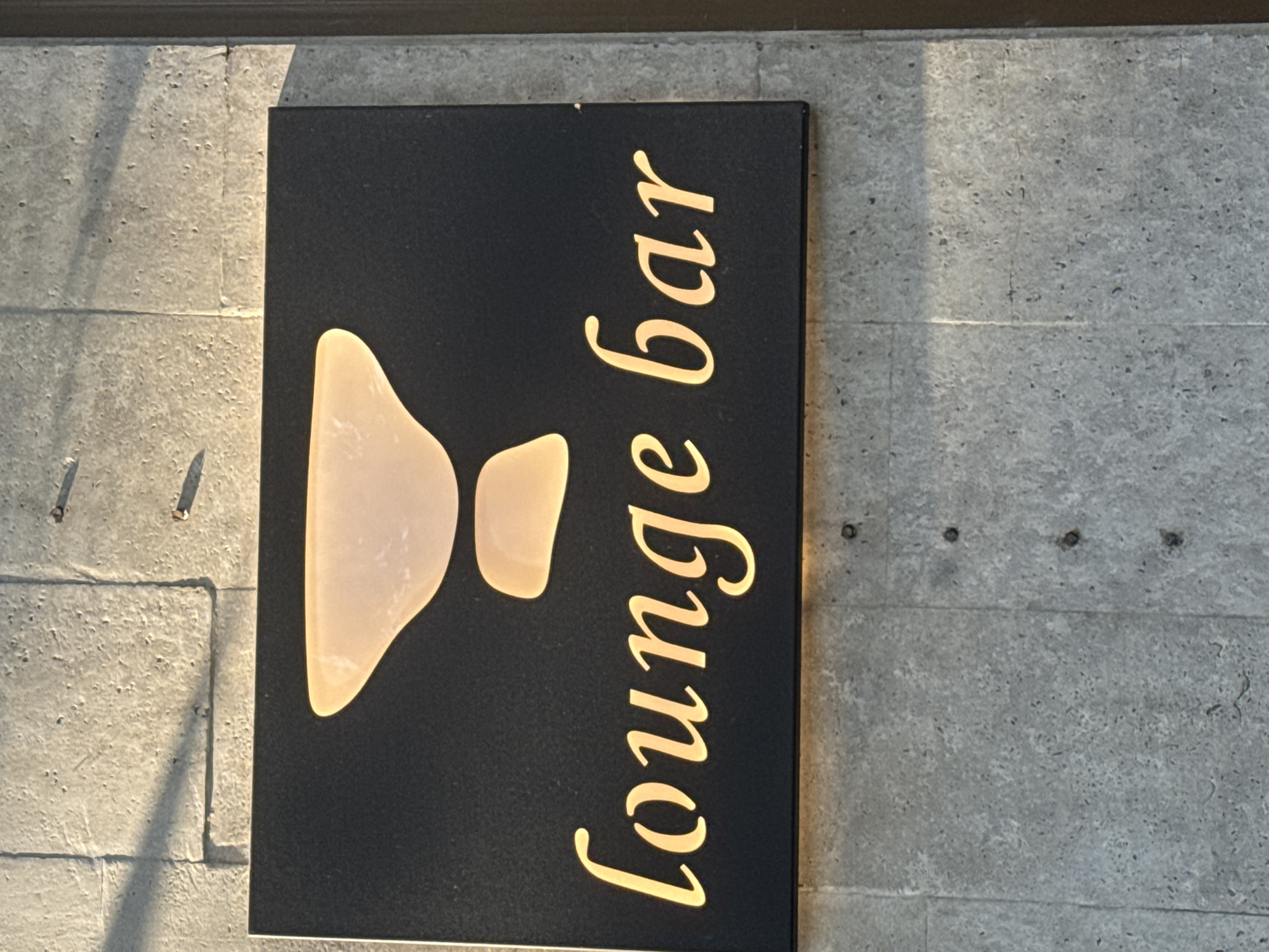 A dark rectangular sign is mounted on a concrete wall. The sign has a stylized illuminated logo at the top, resembling a chalice or goblet with two illuminated sections. Below the logo, the words "lounge bar" are illuminated in a warm yellow font. The concrete wall has visible seams and a rough texture, with some small holes or imperfections. Sunlight casts shadows on the wall, indicating it is outdoors. No people are visible in the image. The location is identified as Bishkek City, Kyrgyzstan.