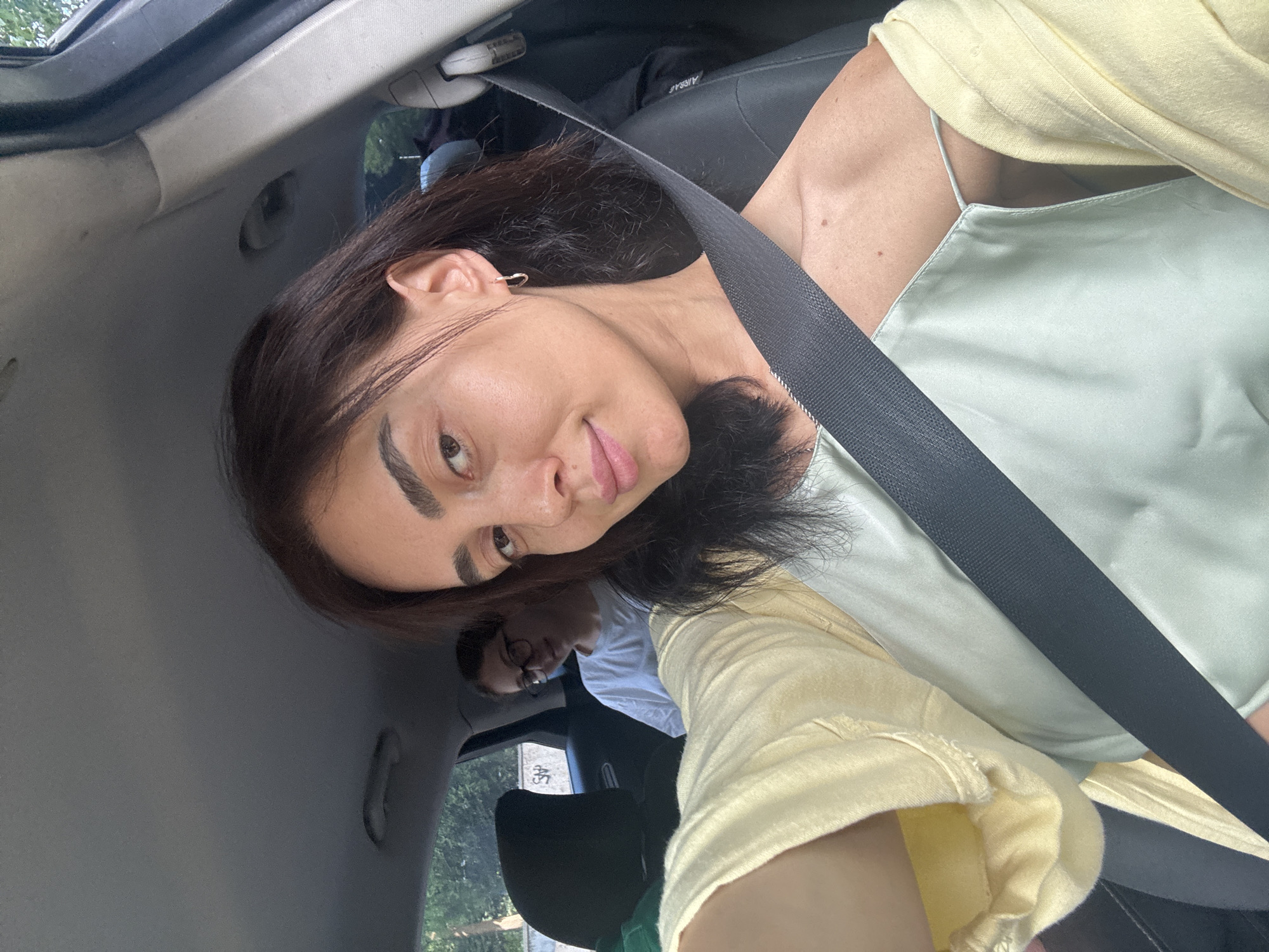 A medium shot captures a woman in the front passenger seat of a car, facing the camera and smiling. She has dark hair, light skin, and is wearing a light yellow cardigan over a pale green camisole. A seatbelt is visible across her chest. In the backseat, a younger person with glasses and a white t-shirt is out of focus. The car interior is visible, including the headrest of the seat in front of the woman and the car's ceiling. The background shows trees and a building with graffiti, suggesting an urban environment. The lighting is natural and appears to be daytime. The location is identified as Tbilisi, Georgia.