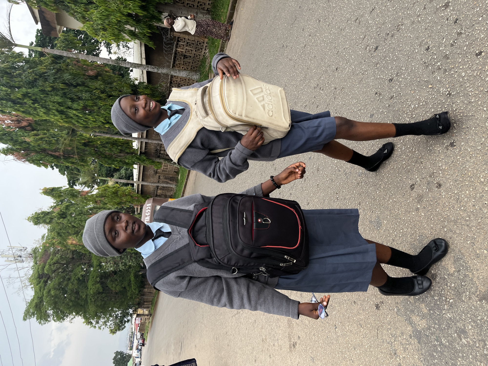 Two young women in school uniforms stand on a paved road. Both wear gray sweaters, blue shirts, gray hats, dark skirts, and black shoes with socks. One carries a black backpack with red trim, and the other has a beige Adidas backpack. The road is flanked by trees, buildings, and a wired fence. Cars are visible in the distance.
