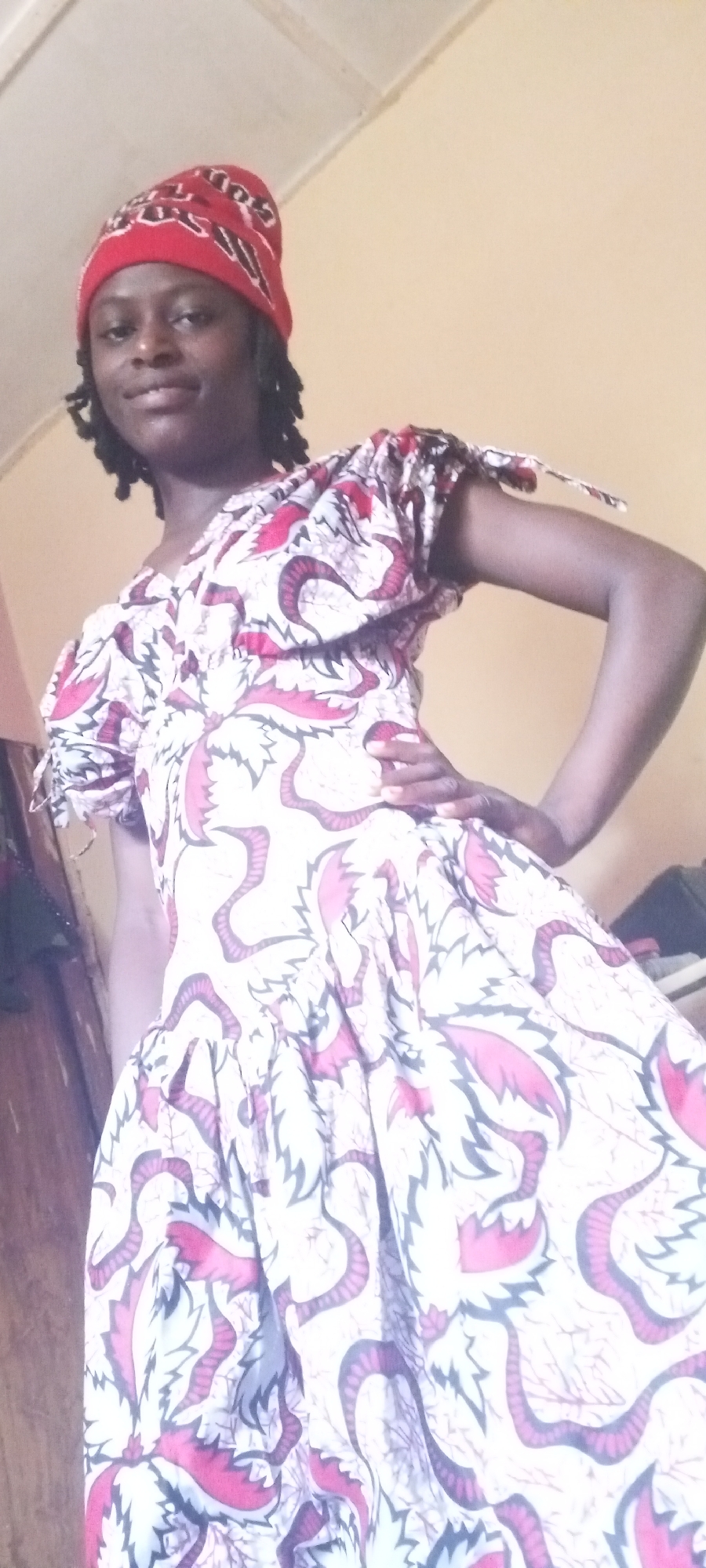 A woman stands indoors in Sabon Tasha, Nigeria. She is wearing a red headscarf and a white dress patterned with red and dark grey designs. The dress has short, puffy sleeves tied at the ends. She has her left hand on her hip and is looking towards the camera with a slight smile. The room is plainly decorated with yellow walls. There is a piece of furniture to the left and other items out of focus in the background on the right.