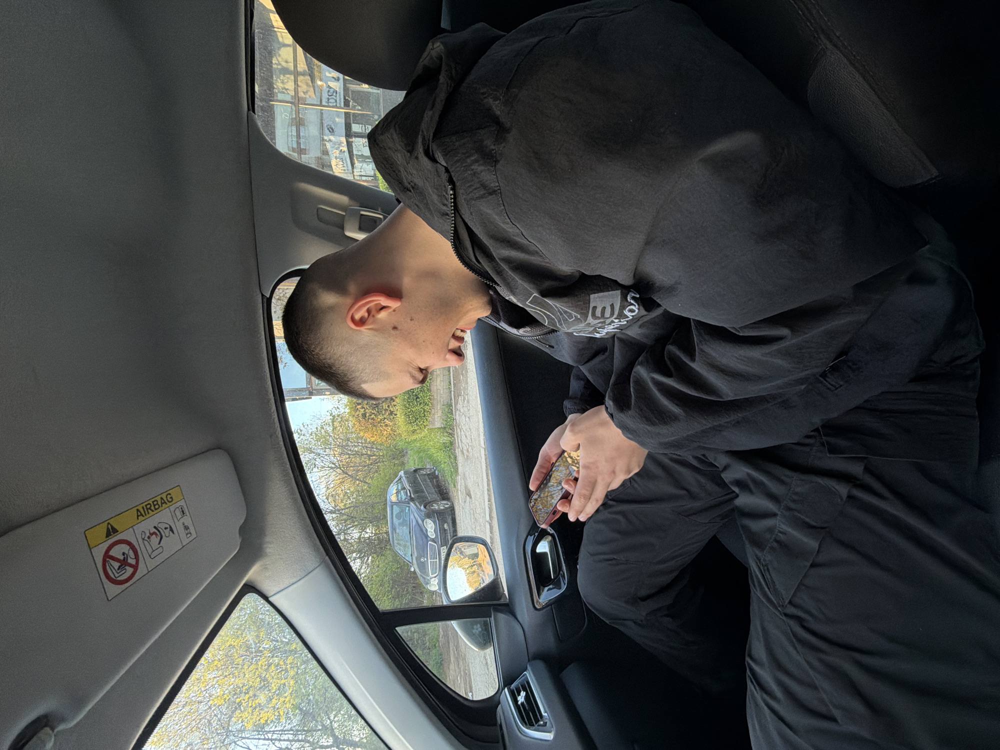 A person sits in the driver's seat of a car. The person is fair-skinned, has short hair, and wears a black jacket and pants. The person is holding a cell phone in their hands. The car is stopped on the side of a street. Outside the car window, another car is parked on the street. Trees line the street, and buildings can be seen in the background. Based on the license plate of the parked vehicle, the location is in Sofia, Bulgaria.