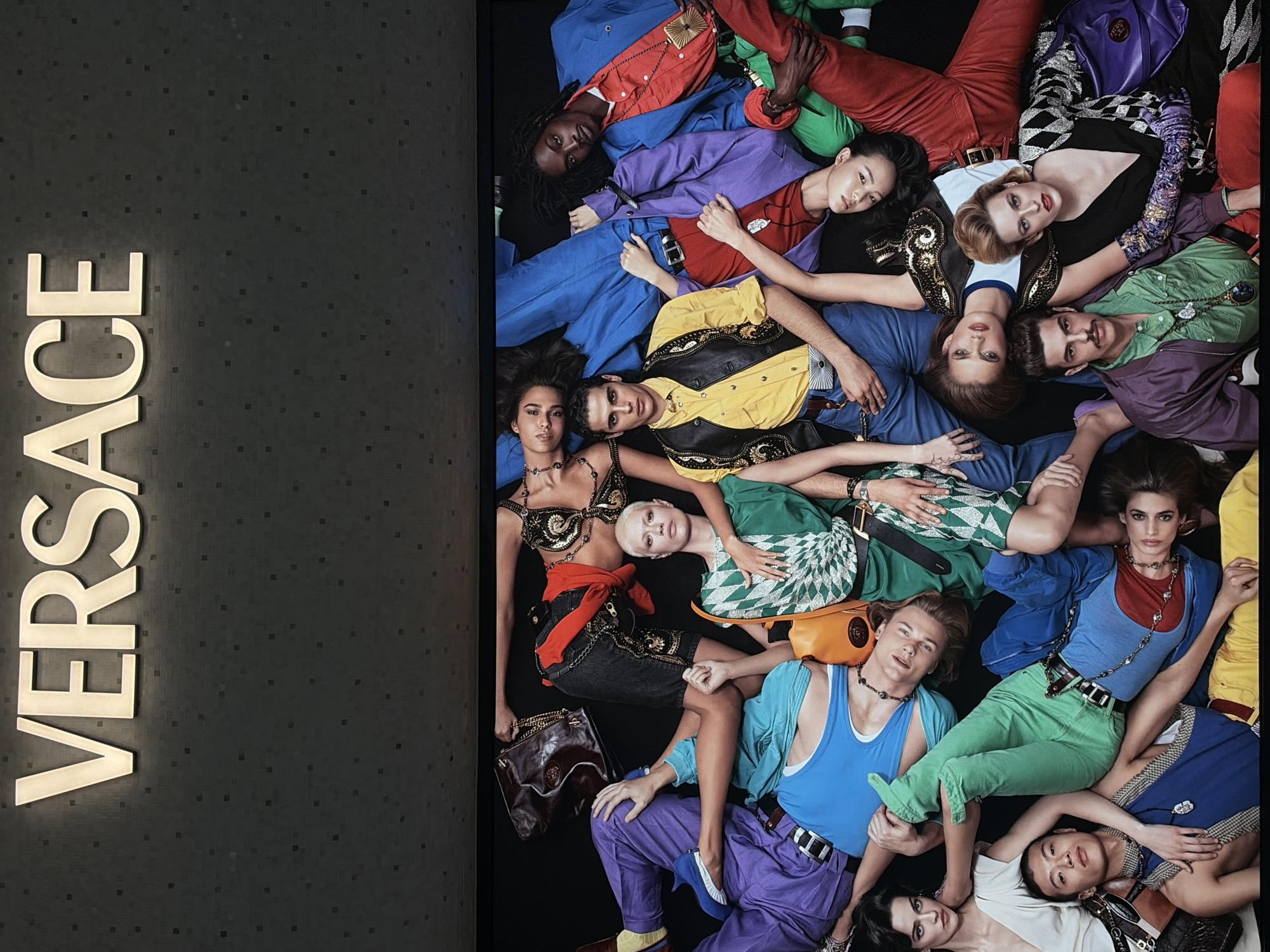 This is a photograph of a Versace advertisement. On the left is a vertical sign with "VERSACE" in large, illuminated gold letters against a dark gray tiled background. To the right of the sign, a group of approximately 15 people are lying in a pile, seemingly on a black background. They are dressed in bright, colorful clothing with various textures and patterns. The clothing appears to be high-fashion, with visible belts, necklaces, and other accessories. The models are diverse in terms of ethnicity and gender, and are posed in a seemingly casual yet styled manner. The photograph suggests a fashion advertisement for Versace. This image was taken at the Iconsiam mall in Bangkok, Thailand.