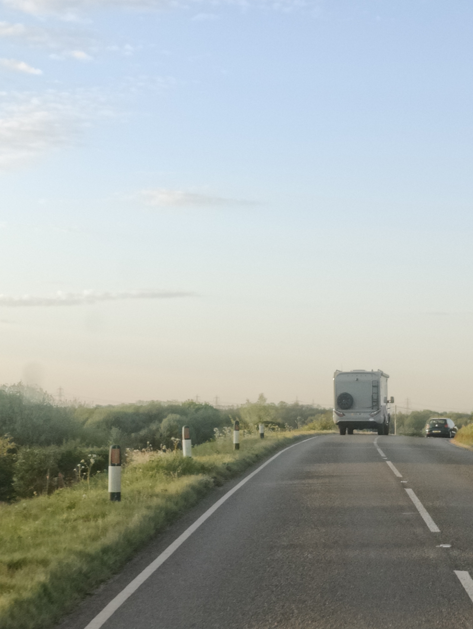 The image depicts a serene, open road scene in East Suffolk, United Kingdom, captured from a moving vehicle. The asphalt road, marked with a solid white line on the left edge and broken white lines in the center, curves gently to the right as it extends into the distance.
Ahead, a large, grey motorhome or RV is prominent, viewed from its rear. It features a spare tire mounted on the back and a ladder on its left side. Further down the road and slightly to the right of the motorhome, a dark-colored car, possibly a hatchback, is also traveling in the same direction.
The left side of the road is bordered by a grassy verge interspersed with white and orange-topped reflective road markers. Beyond the verge, dense green foliage, including bushes and trees, lines the horizon. Faint outlines of power lines or utility poles are visible in the distant haze above the trees.
The sky is a clear, light blue at the top, gradually transitioning to a pale, warm yellow-orange closer to the horizon, suggesting a late afternoon or early evening setting. A few wispy clouds are scattered across the sky. The overall impression is one of peaceful travel through a rural or semi-rural landscape. There is no visible text in the image.
