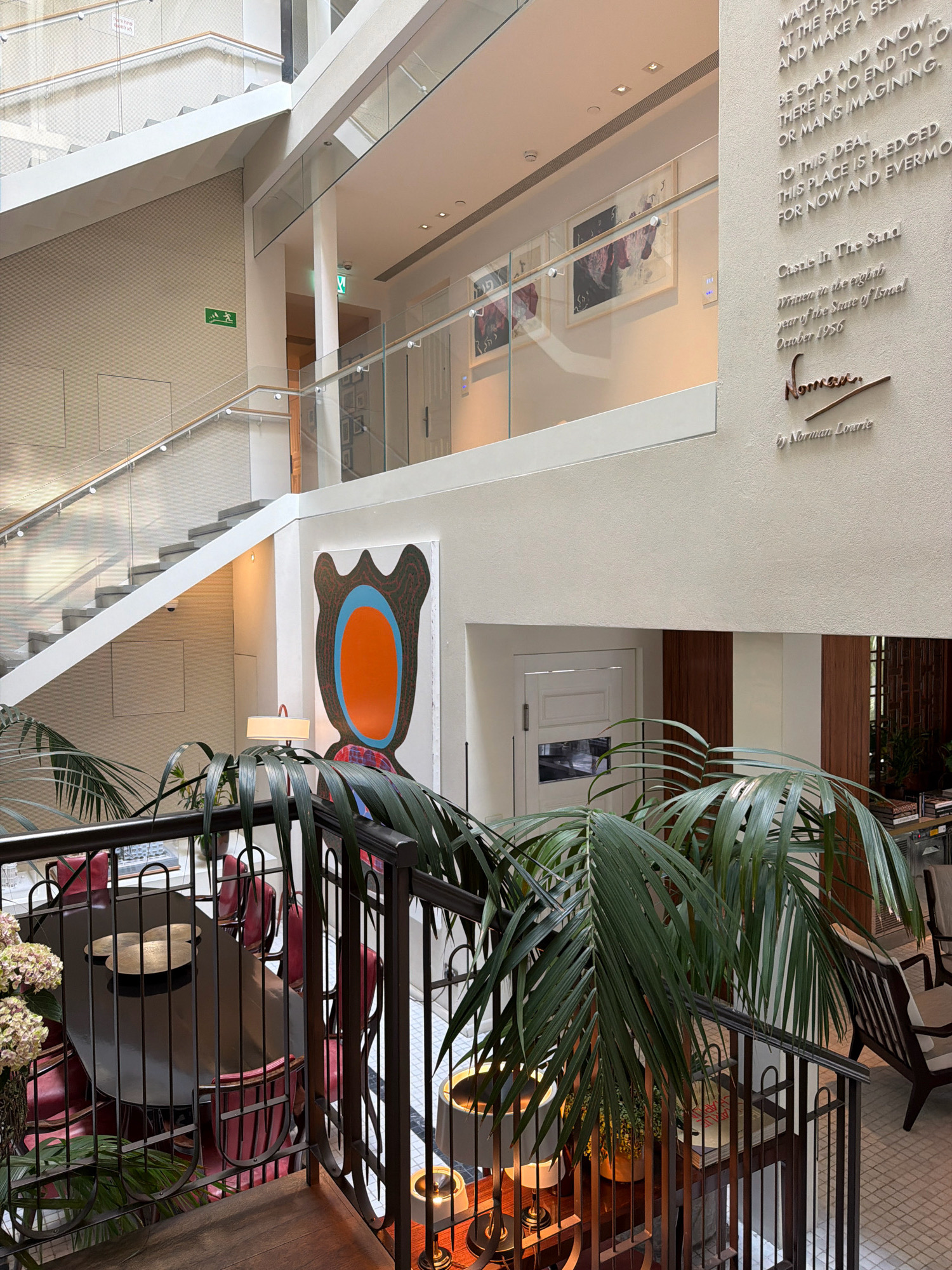 The image shows the interior of a building, likely a hotel lobby or a modern office space, with a focus on a central atrium. A grand staircase with white railings and glass partitions dominates the left side of the frame, leading up to multiple levels. An illuminated green "EXIT" sign is visible on the wall near the staircase.
On the right wall, large white lettering displays text, appearing to be a quote or dedication. Below this text, a plaque with a signature and a name, "Norman Lourie," is mounted.
In the foreground, a lush green palm plant partially obscures a view of a dark oval-shaped table with several red chairs arranged around it. The table appears to be set for a meeting or meal. A table lamp with a white shade is visible near the plant.
Further in the background, several framed artworks hang on the walls. One large, abstract artwork with orange and blue elements is prominent on a white wall. The architectural style suggests a contemporary design with clean lines and natural light flooding the space from above. The overall impression is one of an elegant and well-lit interior.
The text on the wall and the name "Norman Lourie" suggest a connection to Tel-Aviv, Israel, as Norman Lourie was a prominent architect and urban planner in the city. The specific location is likely a building designed or associated with him, possibly the Norman Lourie Building or a space within it.