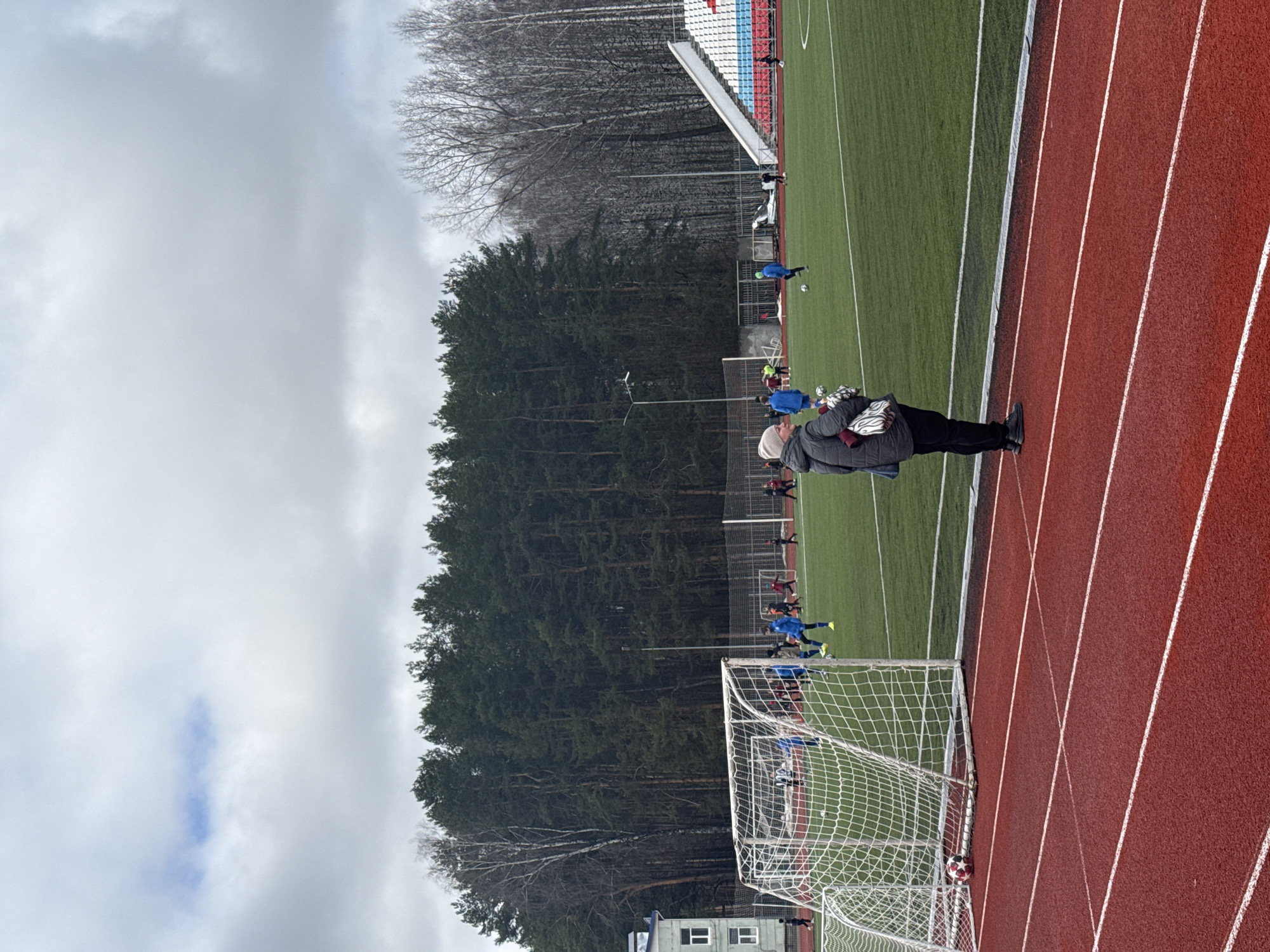 The image depicts a soccer field and track at the stadium in Chaikovsky, Russia. A person stands on the red running track adjacent to the soccer field, observing a soccer practice or game. Several players wearing blue uniforms are on the field. A soccer goal is in the foreground, with a soccer ball nestled in the net. In the background is a dense evergreen forest and a section of stadium seating. The sky is overcast. A building is visible near the goal, likely part of the stadium facilities.