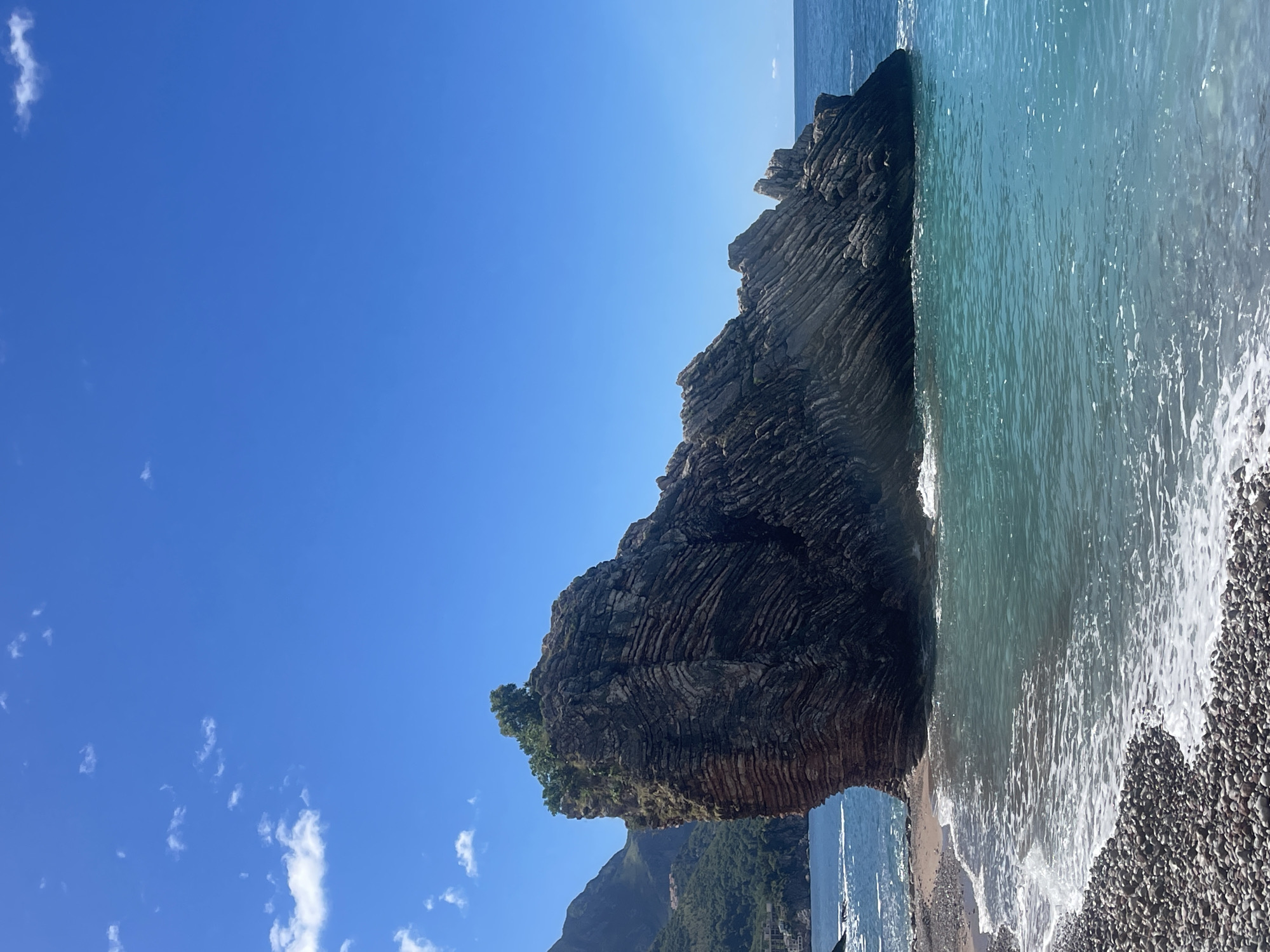 The image shows a rock formation on a beach in Unknown, Unknown. The rock is large and layered, with some vegetation growing on it. The ocean is visible on the right side of the frame, with waves crashing on the beach. The sky is blue with a few scattered clouds. In the background, a mountain is seen covered in greenery. The beach consists of small, rounded stones.