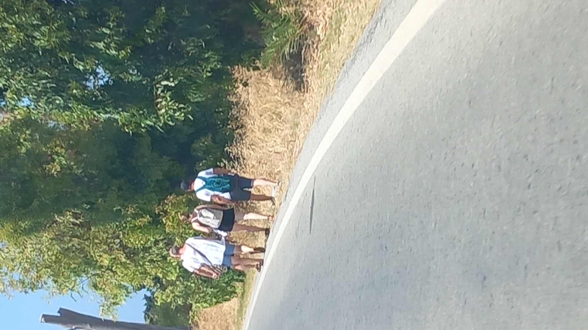 The image shows three people walking along the side of a road in Kos, Greece. The people are walking away from the camera, towards the right side of the frame. The person on the left is wearing a white t-shirt and blue shorts, with a brown bag slung over their shoulder. The middle person is wearing a dark tank top and black shorts, with a light-colored backpack. The person on the right is wearing a white t-shirt, blue shorts, and a teal backpack. They are walking on a dry, grassy shoulder next to an asphalt road. Trees and foliage are visible in the background. The sky is clear and blue.