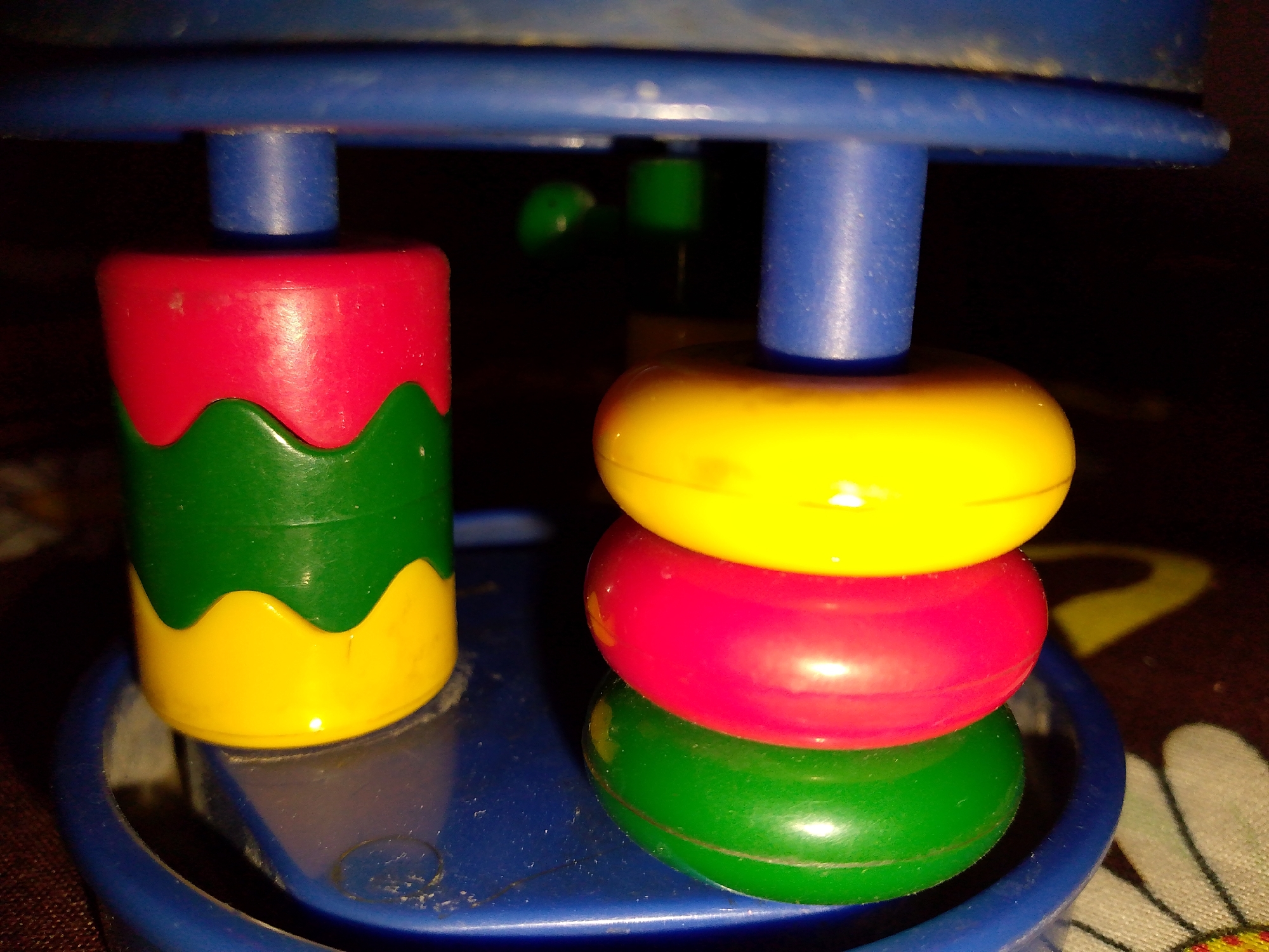 The image shows a close-up of a toy. In the foreground, three colorful stacking rings are piled on top of each other: yellow, red, and green. To the left of the rings, a red and green cylindrical toy with a wavy pattern is visible. The toys are resting on a blue base. The background is dark and appears to be a patterned surface. The lighting is dim, creating shadows. The location is unknown, and no people are present in the image.