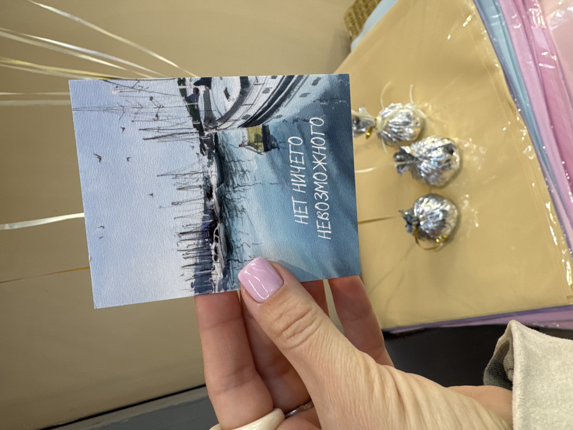A postcard featuring a maritime scene is held in a person's hand. The scene appears to be a harbor with numerous sailboats. The postcard's text, written in Russian, reads "НЕТ НИЧЕГО НЕВОЗМОЖНОГО", which translates to "NOTHING IS IMPOSSIBLE". The woman has a light pink nail polish on her fingernail. Behind the postcard, there are foil-wrapped candies and light-colored wrapping materials, suggesting a possible gift or present. The background includes a beige wall, and a glimpse of a gray fabric is visible in the lower right corner. Given that the text is in Russian and the image evokes maritime imagery, it is plausible that the location is in Sochi, Russia, as Sochi is a port city on the Black Sea.