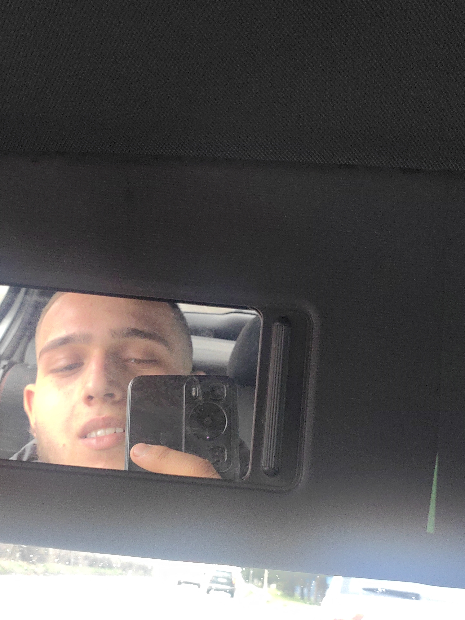 The image shows a man taking a selfie in a car mirror. The man has short hair and is holding a black smartphone. The mirror is part of the car's sun visor. In the reflection, a road with cars and greenery can be seen outside the car. The location is unknown, and it is not possible to verify that the image was taken in Sofia, Bulgaria, from the given visual data.