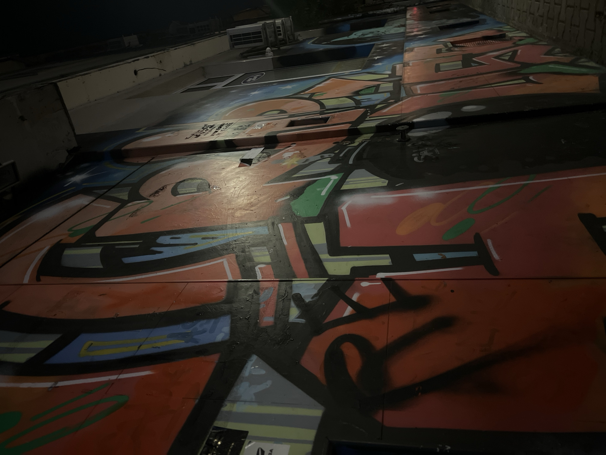 The image shows a graffiti-covered wall of a building at night. The graffiti features bold, colorful letters and designs in shades of orange, red, blue, green, and yellow, outlined in black. Some areas of the wall appear to be made of corrugated metal. There are several windows and what looks like an air conditioning unit visible on the building. The ground in front of the wall is paved with brickwork. The overall impression is of an urban alleyway or street scene at night, illuminated by artificial light. The graffiti artist's name "REDF" is partially visible on the left side of the wall. While no specific Sofia, Bulgaria landmarks are explicitly identifiable, the style of graffiti and urban setting are consistent with many cities.