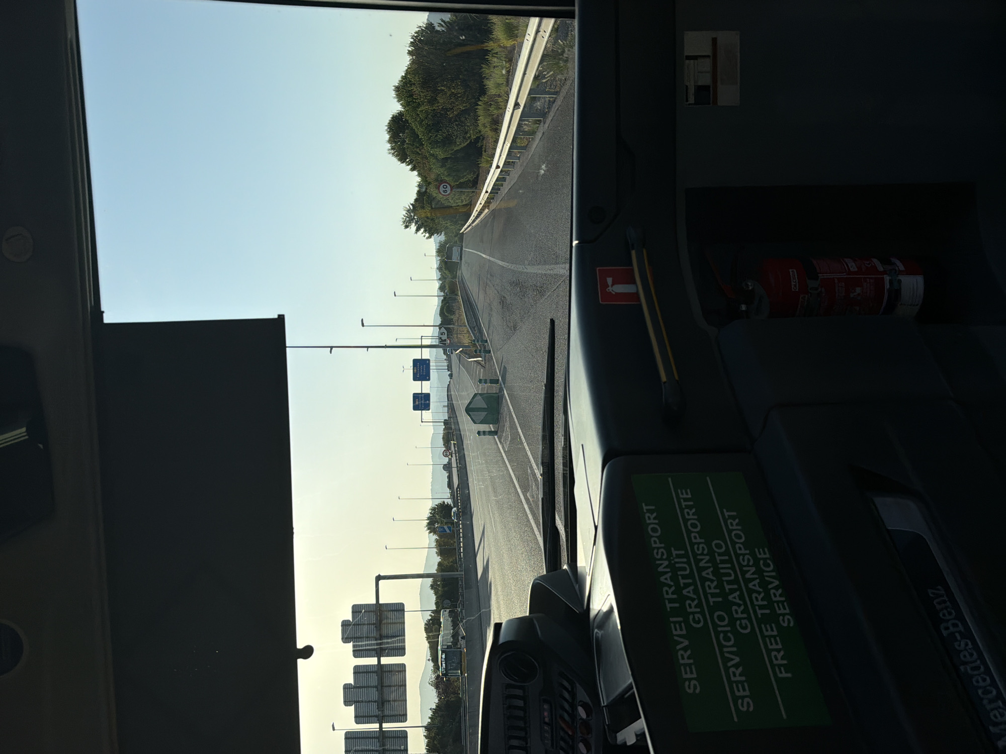 This is a first-person view from inside a Mercedes-Benz bus, likely in El Prat de Llobregat, Spain, as indicated by the context of the other images from this set. The bus is traveling on a highway with multiple lanes. In the foreground, the dashboard of the bus is visible, featuring various controls and a green sticker with text in Portuguese, Spanish, and English stating "SERVEI TRANSPORT GRATUIT SERVICIO TRANSPORTE GRATUITO FREE TRANSPORT SERVICE". A fire extinguisher is mounted on the right side of the dashboard. The view through the windshield shows a clear blue sky and a highway stretching into the distance. Several highway signs and streetlights line the road. In the mid-ground, another bus is visible on an adjacent road, and further down the highway, trees and vegetation are present. The sun is low in the sky, suggesting it is either early morning or late afternoon. The overall scene depicts a journey on a sunny day, with a focus on the interior of the bus and the road ahead.