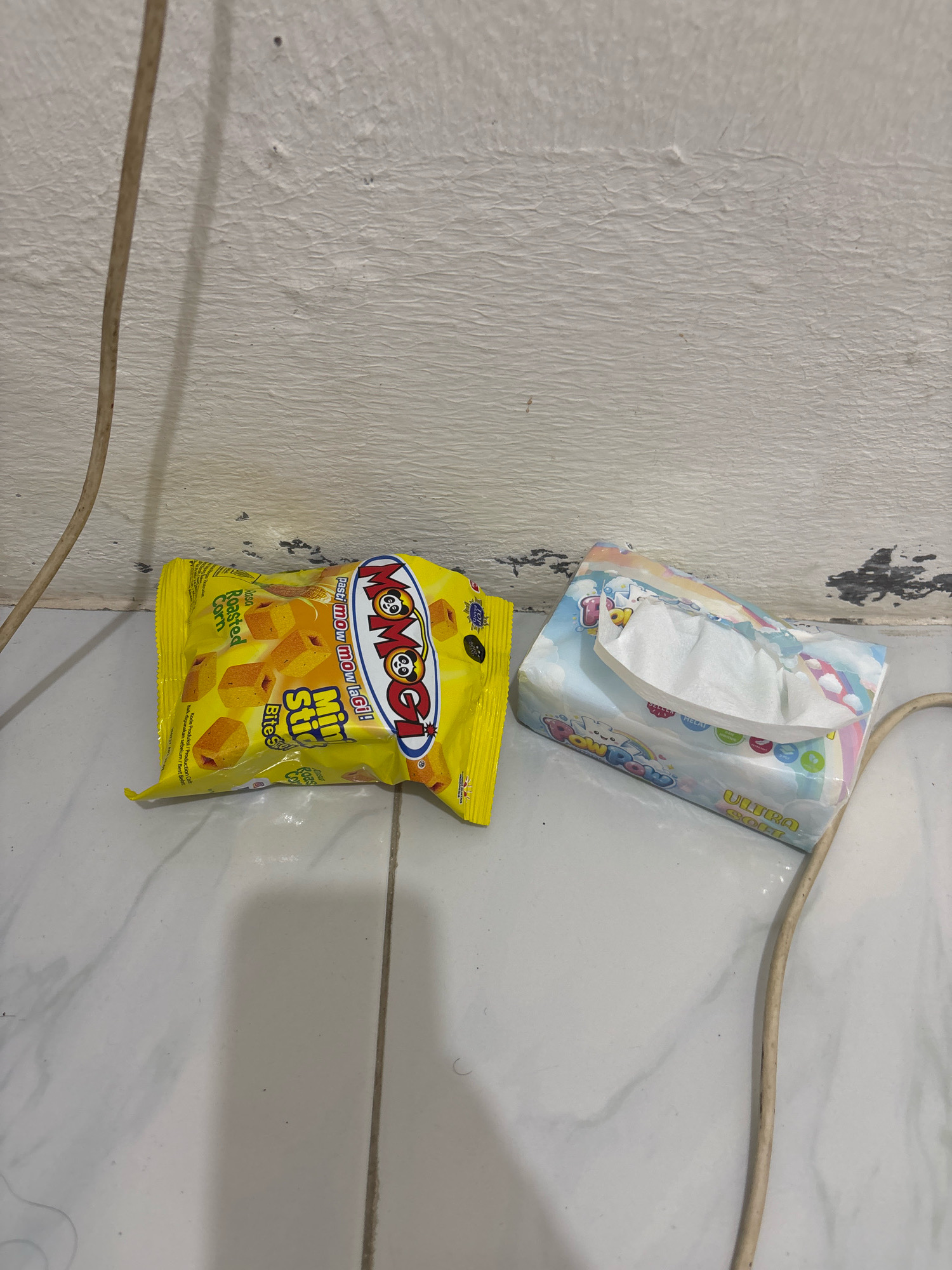 The image shows two items on a tiled floor next to a white, textured wall. On the left is a yellow snack bag with "Momogi Mini Stick Bites" and "Roasted Corn" printed on it. To the right of the snack bag is a package of tissues with a colorful design featuring clouds and a rainbow. A beige electrical cord runs from the upper left corner of the image down towards the bottom right, with a shadow cast by another object not fully visible. The environment appears to be indoors, possibly a home or a shop, in Wanajaya, Indonesia.