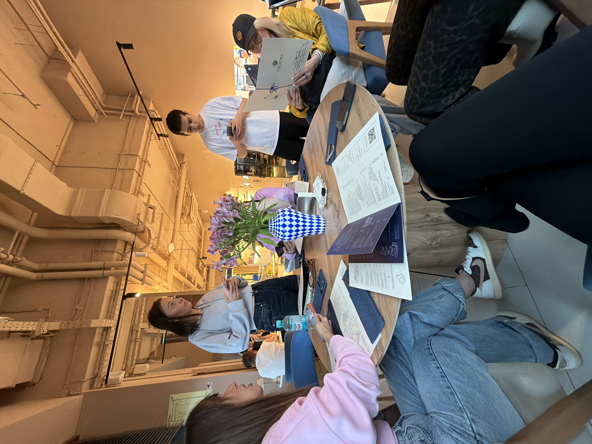 The scene is indoors, in what appears to be a restaurant. There is a group of people, mostly women, gathered around a circular wooden table. They are looking at menus and interacting with each other, suggesting they are about to order food.
The environment includes exposed ductwork and industrial-style lighting fixtures on the ceiling. The table is adorned with menus, eyeglasses, water bottles, a vase of flowers in a blue and white checkered container.
The location is in Khimki, Russia.