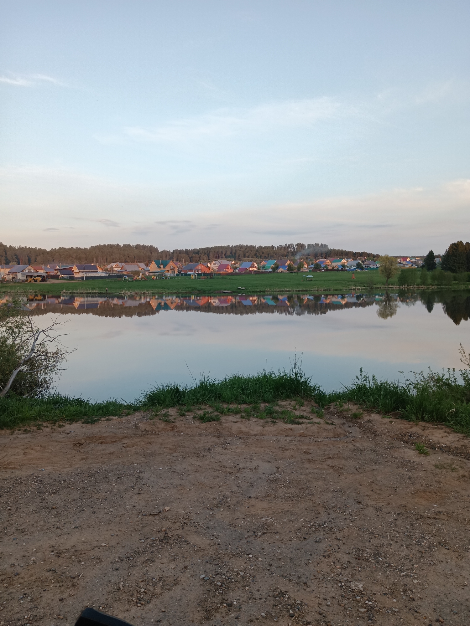 The image shows a serene scene in Фоки, Russia. A body of water reflects the houses and trees lining the opposite bank, creating a mirror-like effect. The water surface is calm. The houses have a variety of roof colors. In the foreground, there is a dirt or gravel area with some grass and vegetation growing near the water's edge. The sky is light blue with some clouds. The light suggests it is either early morning or late afternoon. No people are visible in the shot.