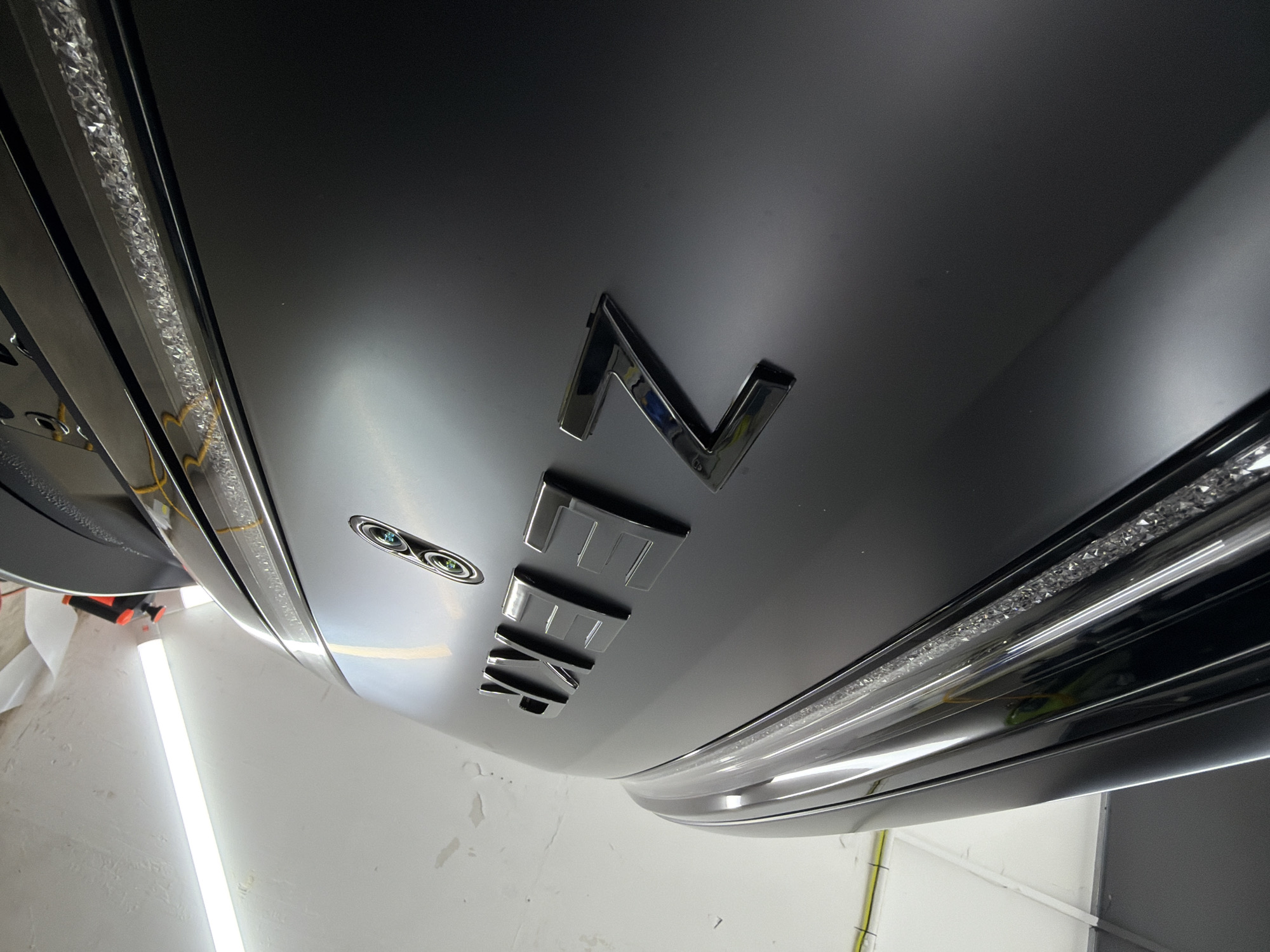 A close-up, downward-facing perspective shows a portion of a car's matte grey trunk with "ZENIT" spelled out in chrome block letters. The car also has LED lights installed. The location is likely an automobile service or detailing shop. The location is unknown.