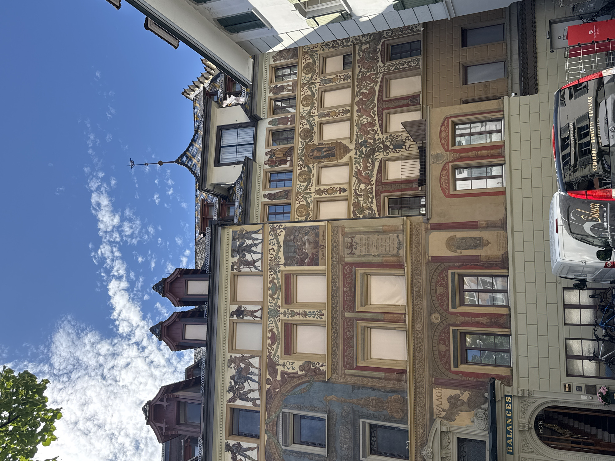 The image shows a detailed facade of buildings in Lucerne, Switzerland, featuring ornate murals and architectural elements under a bright blue sky with scattered clouds. The buildings are decorated with historical scenes and figures painted directly onto the walls. One building displays a mural depicting armored figures in the upper register, and elaborate friezes and figures in the lower registers. Another section shows people in historical attire and scenes that appear to be religious or mythological in nature. The building to the left has a sign that reads "BALANCES HOTEL." A street level entrance to the hotel is visible, as well as bicycles parked outside. A white van and a black van with text indicating "HOTEL DES BALANCES," "SEMINAIRE - BANQUETTE - CATERING," and a website address are parked in front of the buildings. The architectural style suggests a historic European city. The scene is static, with no discernible human activity beyond the parked vehicles and bicycles. The lighting indicates it is daytime, likely late morning or early afternoon, given the shadows.