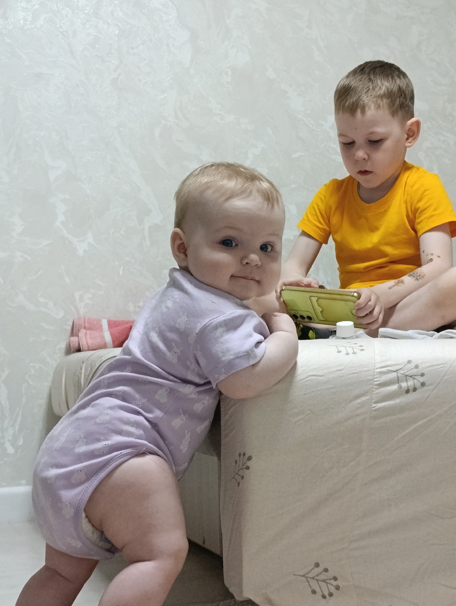 The image captures two young children in an indoor setting in what appears to be a home. The foreground features a baby in a light purple bodysuit patterned with rabbits, leaning against a beige surface possibly a bed. The baby is fair-skinned with blonde hair and blue eyes, looking directly at the camera.
In the background, a slightly older boy wearing a yellow t-shirt is sitting cross-legged on the bed. He is fair-skinned with short brown hair, focused on a yellow smartphone or device. There are also what appear to be temporary tattoos on his left arm. A small white container is located near the phone.
The background includes a light-colored wall with a textured or patterned finish. A folded pink and white striped towel is placed behind the baby.
Based on metadata, the image was taken in the city of Chaikovsky, Perm Krai, Russia.