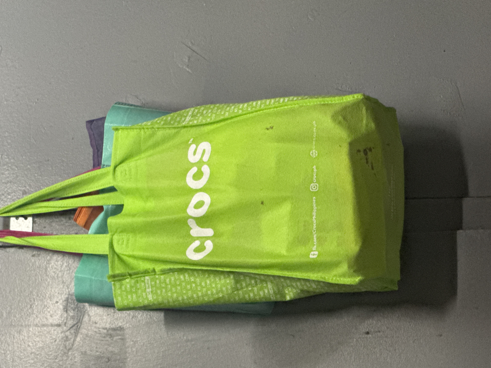 The image features a collection of reusable shopping bags, with a bright lime green Crocs branded bag prominently displayed in the foreground. The bags are set against a plain, textured grey surface, which appears to be a concrete floor or a painted wall, suggesting an indoor or covered setting. The lighting is even and somewhat dim, without harsh shadows.
The main Crocs bag has "crocs" printed in large white letters across its front. Smaller white text is visible vertically along the right side of the bag, providing brand contact information: "fb.com/CrocsPhilippines," "@crocsph," and "www.crocs.com.ph." These details explicitly link the item to the Crocs brand within the Philippines, consistent with the location context of Carmen, Philippines. The bag shows some minor dark stains or smudges on its surface, indicating prior use.
Behind the vibrant green Crocs bag, several other folded reusable bags are visible in shades of teal, purple, and orange. The bags are likely stored together. No specific activity or event is depicted; it is a static image of stored objects. There are no people in the frame, and no particular emotions are conveyed by the scene. The time of day is not discernible, but the lighting suggests an indoor environment or overcast conditions.