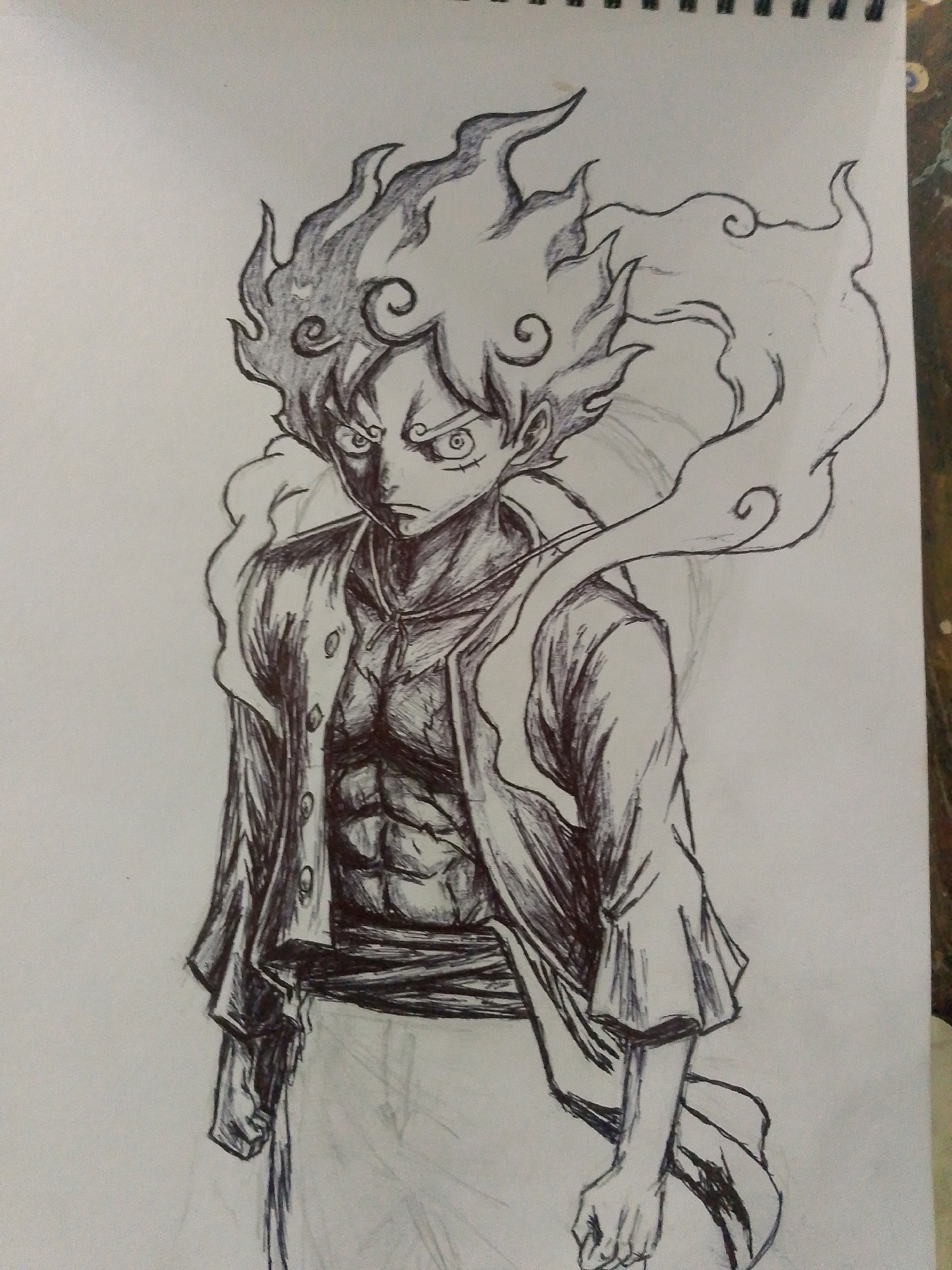 A black and white pen drawing depicts a muscular male anime character with spiky, flame-like hair. He wears an open, unbuttoned shirt over a visible toned chest and abdomen, and a sash around his waist. The character has an intense expression with sharp eyes. The drawing is on a piece of paper, likely from a sketchbook, with the spiral binding visible at the top. The background is plain white. No location information or context about Sitarampuram, India can be inferred from the image.