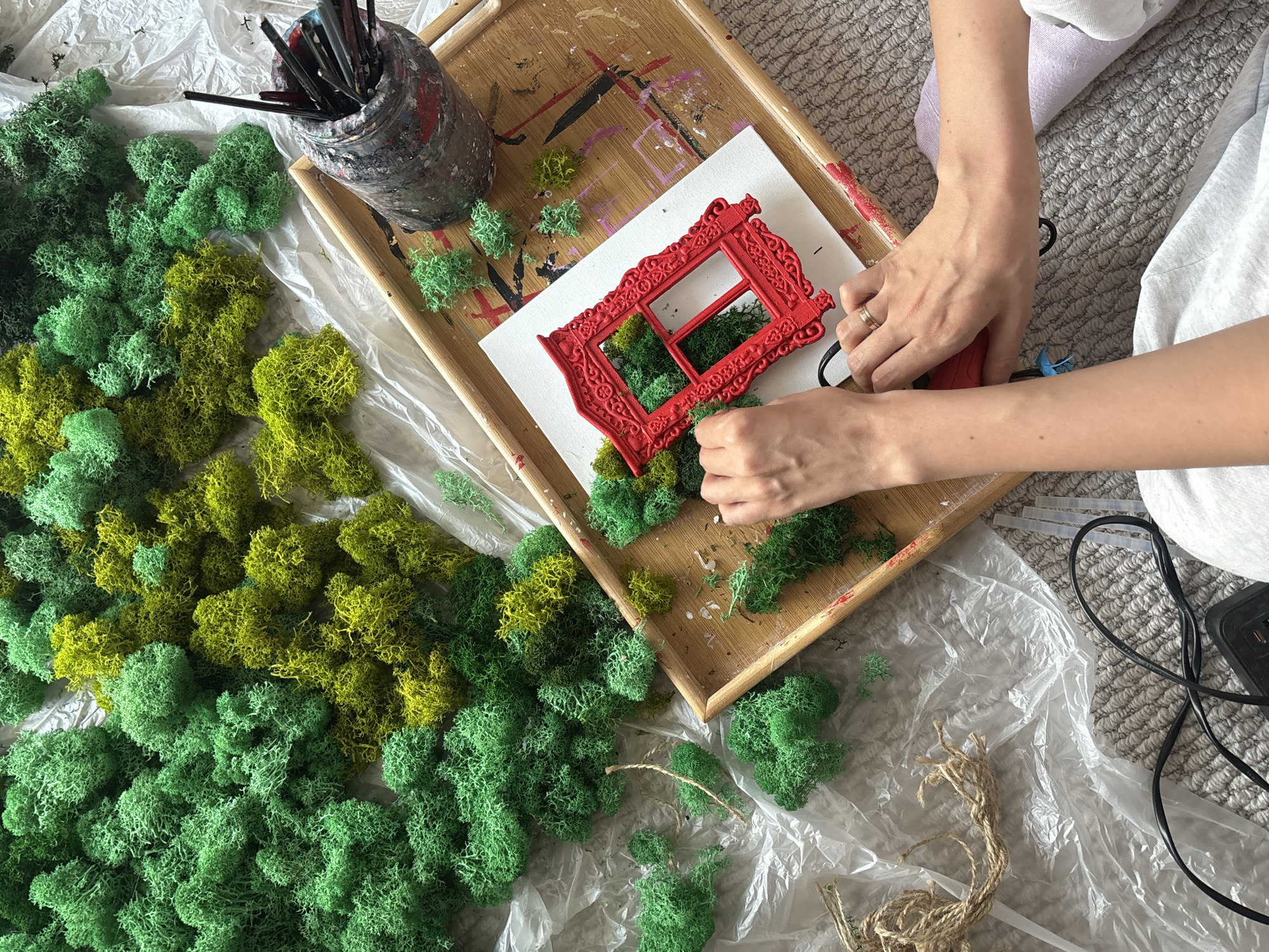 The image shows a person crafting indoors. The individual is using glue to affix green and yellow moss to a white canvas with a red decorative frame on it. Crafting supplies are scattered around, including a tray, paintbrushes in a container, string, and a plastic sheet covering the carpet. The location is unknown.