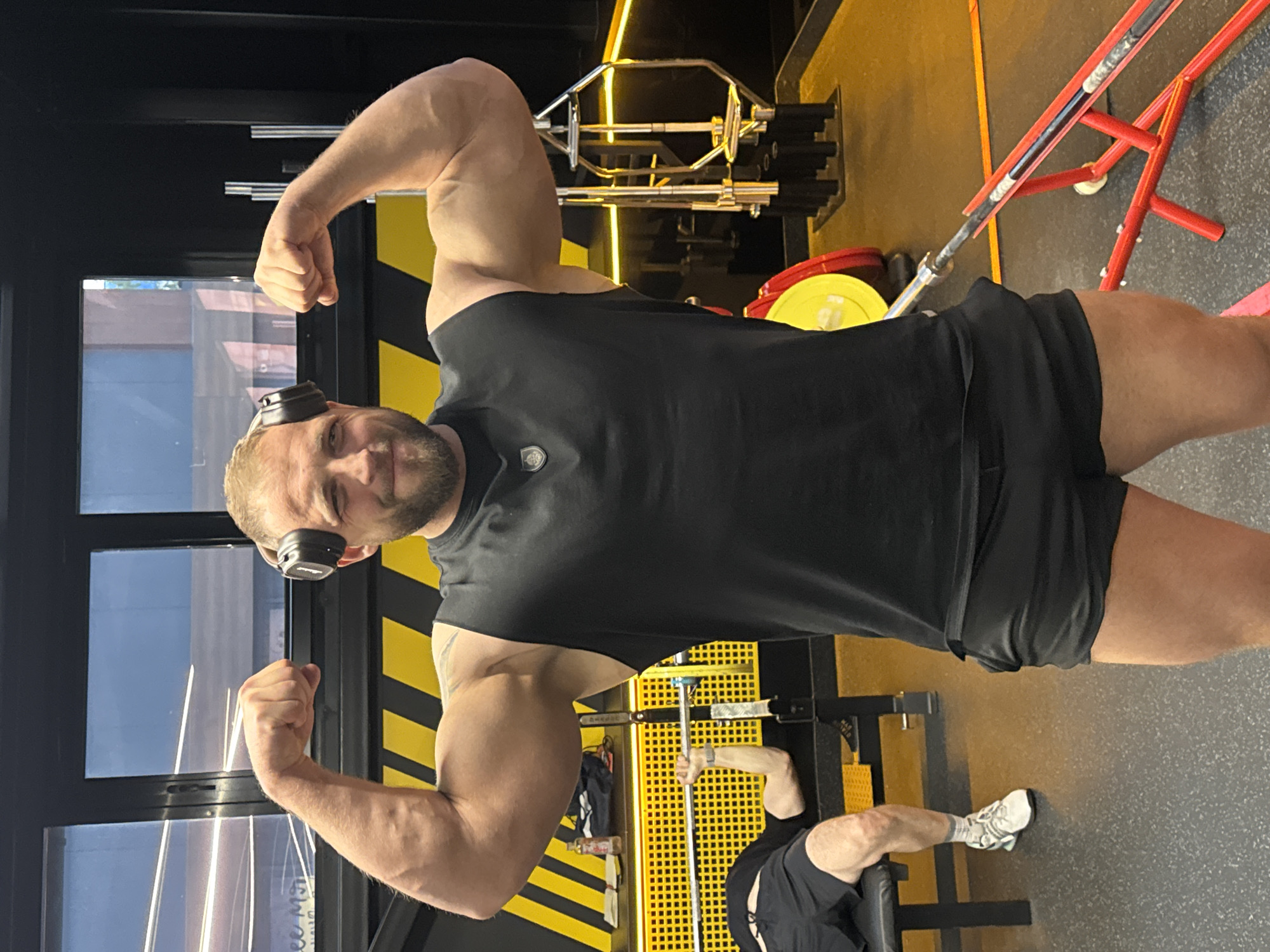 The image displays a muscular man posing inside a modern gym, captured in a portrait orientation but appearing rotated 90 degrees counter-clockwise. The man is the primary subject, flexing both biceps towards the camera with a confident expression and a slight smile. He has a beard and is wearing black over-ear headphones, a black sleeveless top, and black shorts. A small, dark emblem is visible on his chest.
The setting is a contemporary gym characterized by dark walls and a grey, textured floor. Bright yellow strip lighting runs along the walls and ceiling, illuminating the space. Various gym equipment is visible, including a hexagonal barbell on a rack, other barbells, and yellow and red weight plates. A red metal frame, likely part of a squat rack or similar apparatus, stands in the background.
Through a large window on the left side of the frame, outdoor reflections and faint, indecipherable text can be seen. In the lower background, another person's legs and feet, wearing white sneakers and grey socks, are visible as they appear to be seated on a piece of gym equipment, possibly engaged in an exercise. Black and yellow striped patterns adorn sections of the walls and equipment, contributing to the gym's distinct aesthetic. The overall activity depicted is a posed moment, likely a progress picture or a display of strength, within an active gym environment.