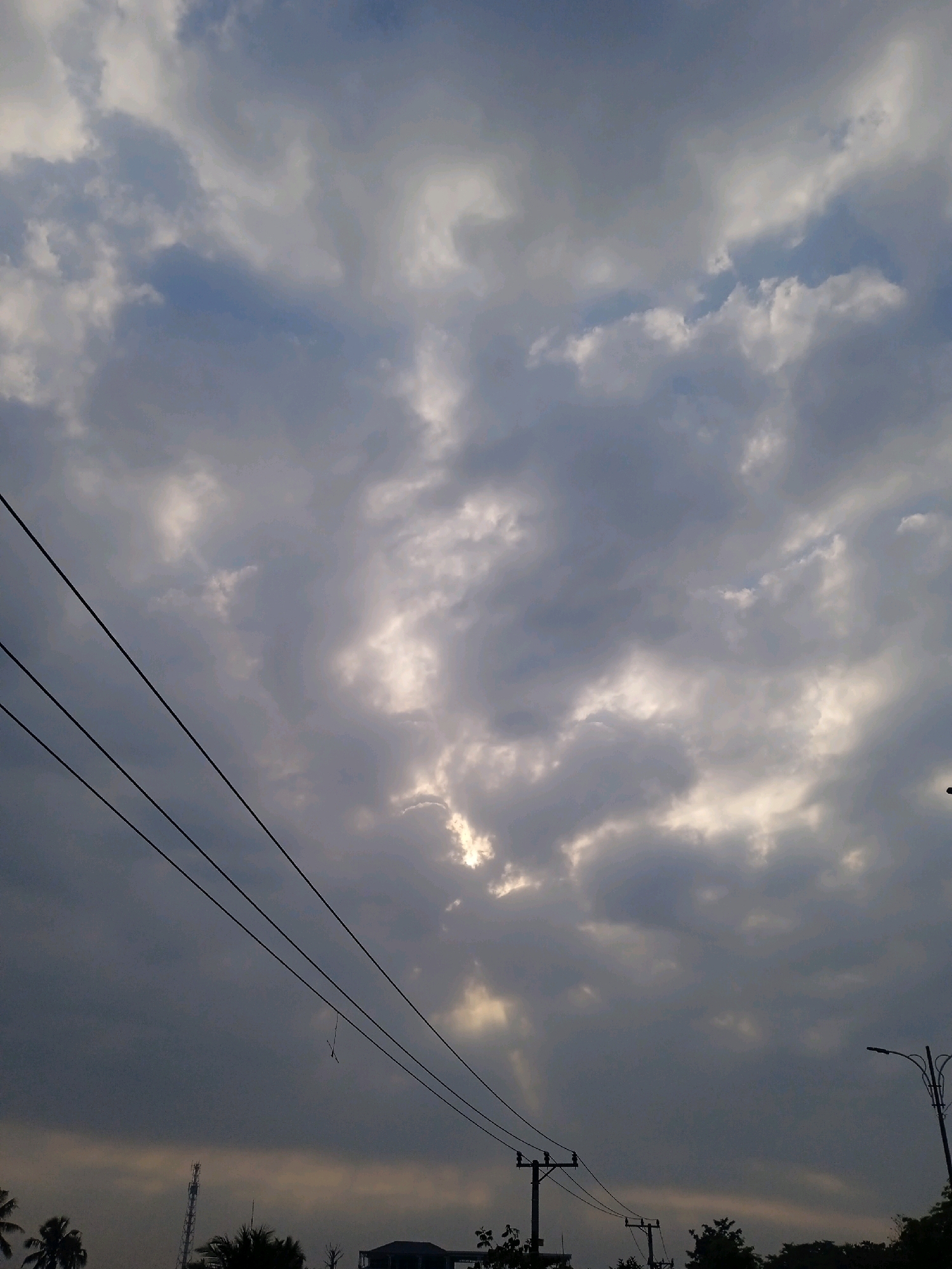 The image captures a cloudy sky with dramatic light rays piercing through. Three parallel power lines stretch diagonally from the left side of the frame towards the upper right. In the lower portion of the image, silhouettes of trees and utility poles are visible, suggesting an urban or suburban environment. A tall communication tower can be seen on the left, and a street light on the right. The overall impression is that of a sky before or after a rain shower, with the sun intermittently breaking through the clouds. The location is identified as Banjarmasin, Indonesia, implying this scene is a typical view of the sky in that region.